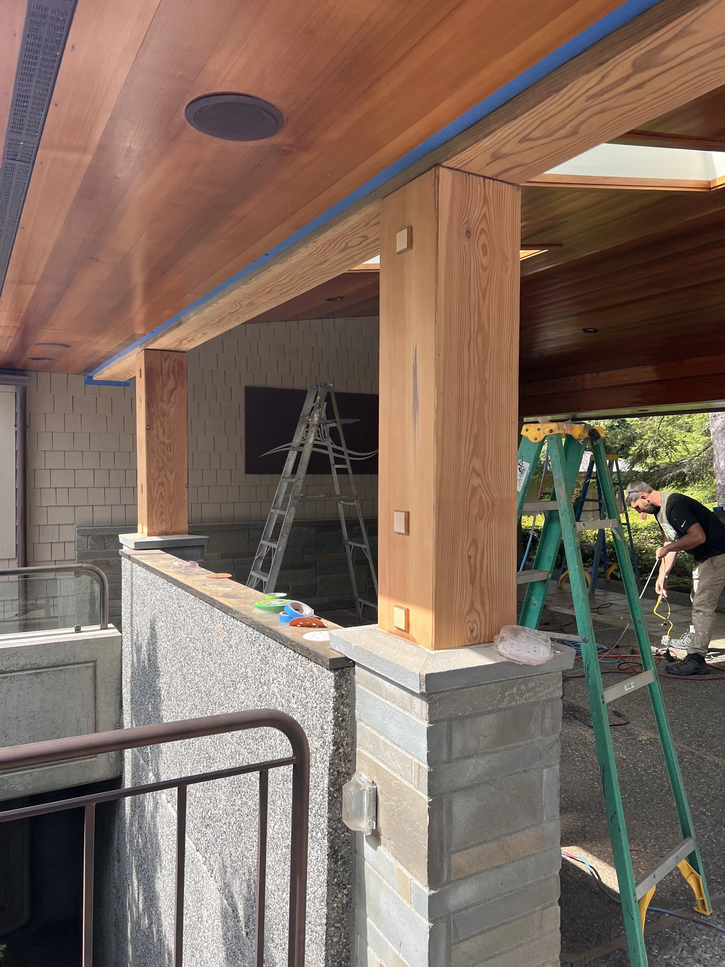 Refinishing wooden beams on a porch. There are ladders, tools, and building materials visible, with a brick wall and greenery in the background.