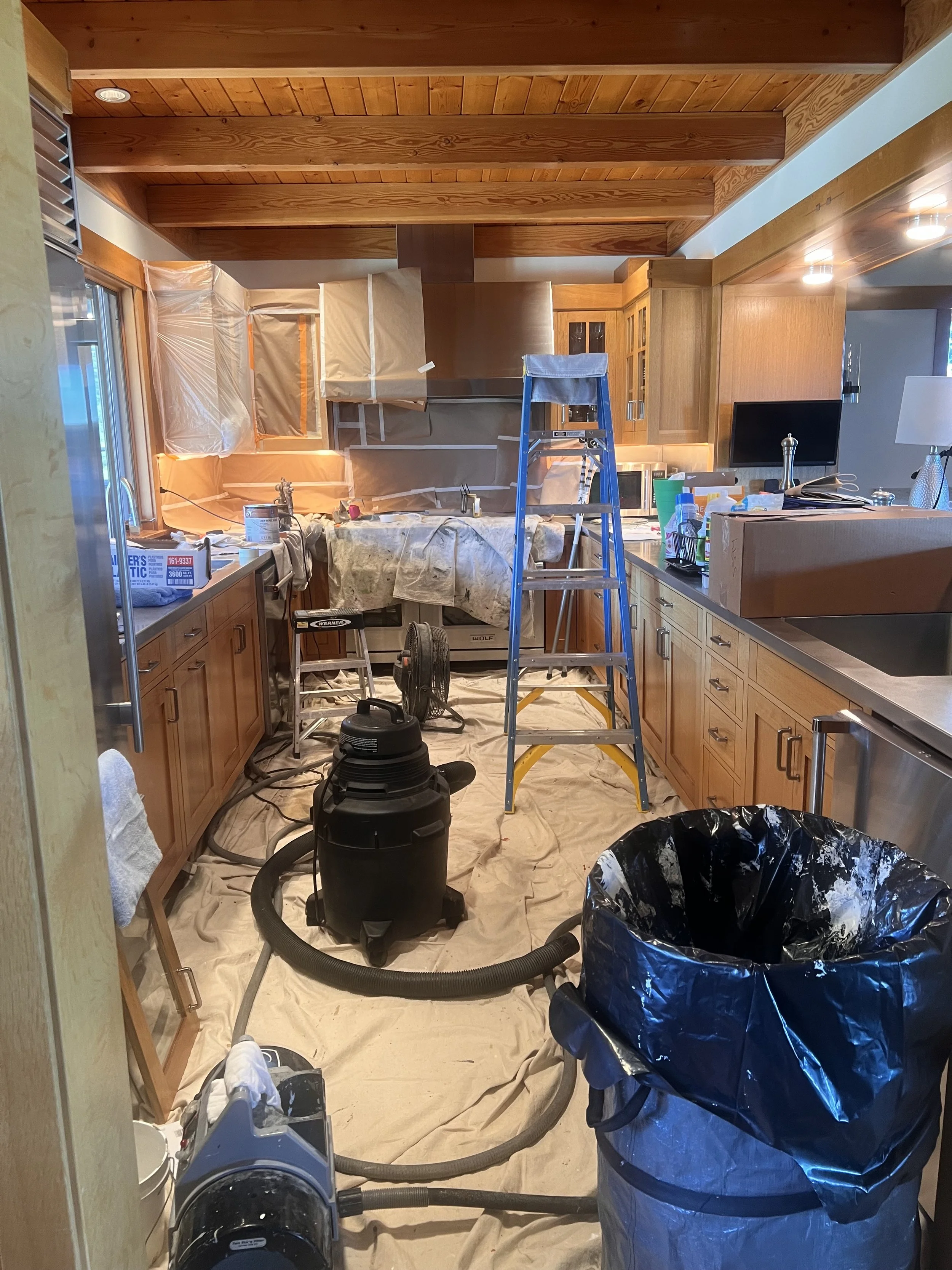 A kitchen under renovation with wooden cabinets, a ladder, a black vacuum cleaner, cleaning supplies, and covered windows. There are construction materials and tools on the countertops and paper covering the floor.