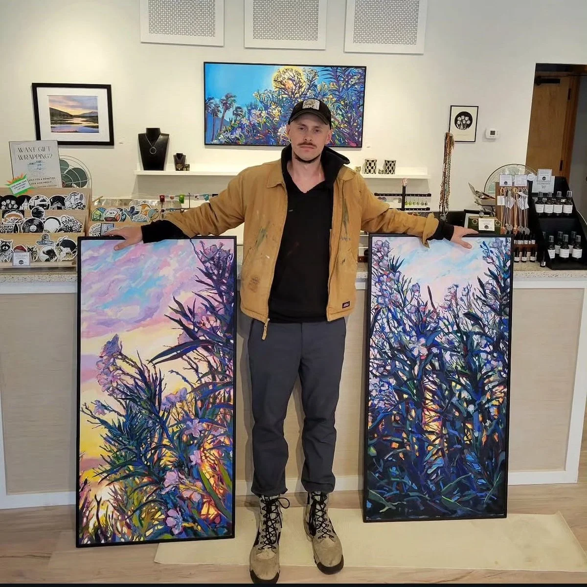 Benjamin standing in art gallery holding two large framed landscape paintings featuring trees and colorful skies.