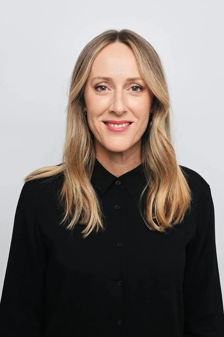 A woman with long, wavy blonde hair smiling, wearing a black button-up shirt, standing against a plain light gray background.
