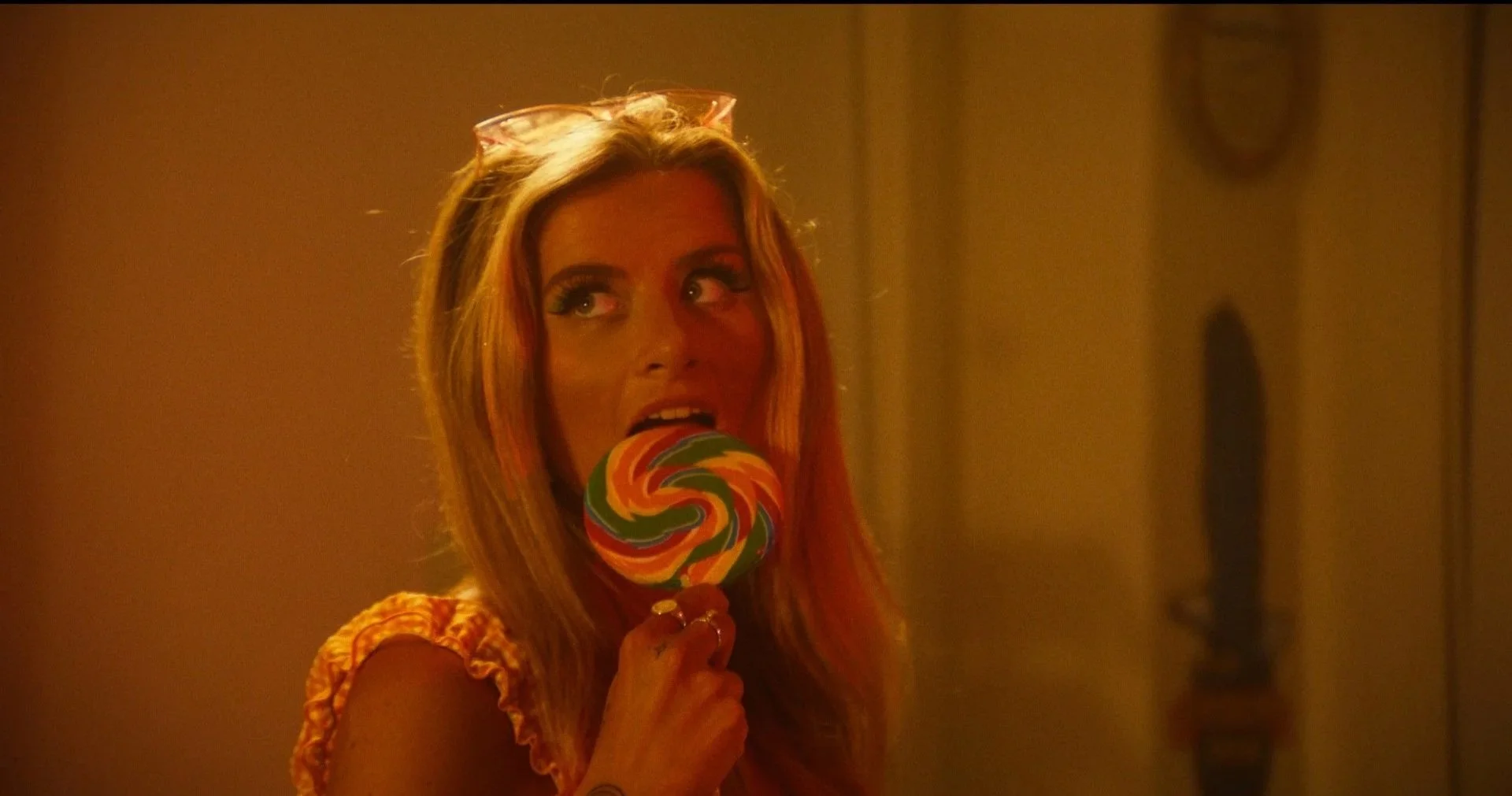 A woman with blonde hair and colorful makeup is biting a large multicolored swirl lollipop, standing in a dimly lit room with warm lighting.