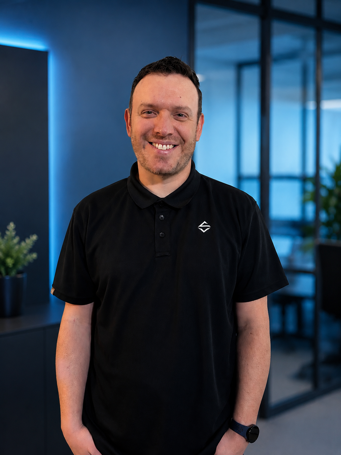 Smiling man in black polo shirt standing in modern office with blue lighting and glass windows.