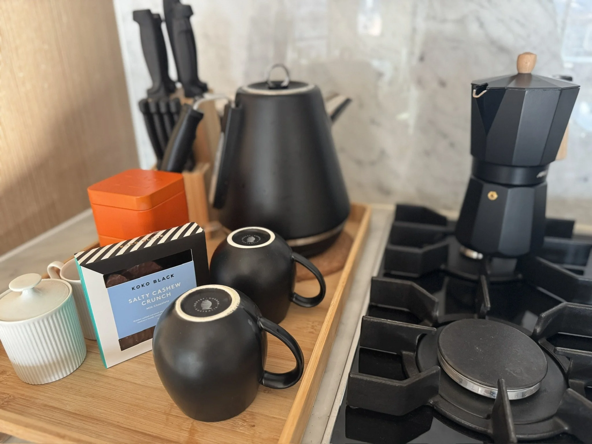 Kitchen countertop with black electric kettle, black coffee grinder, orange container, and cups, next to stove with black burners and grates.