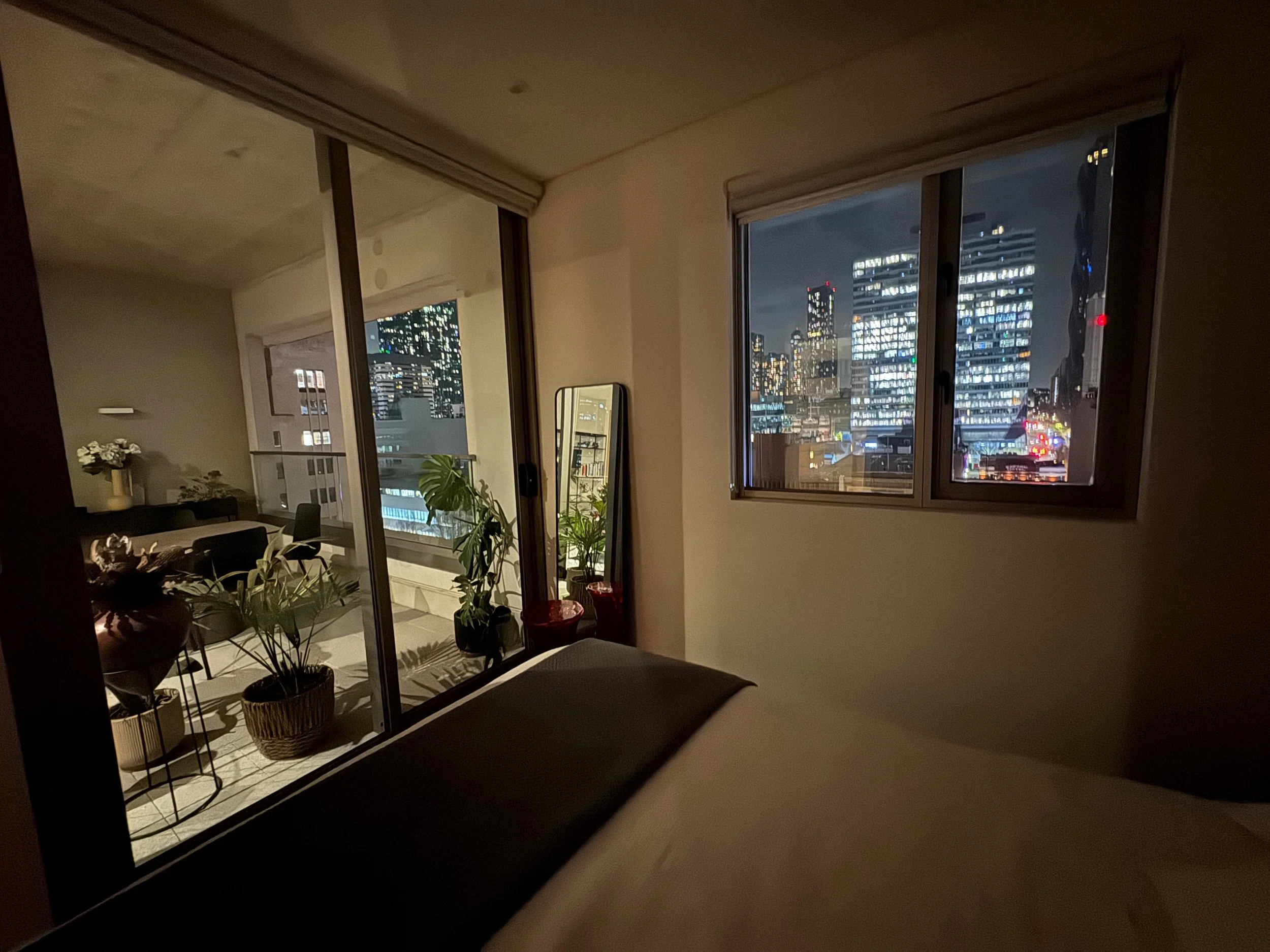 Nighttime cityscape view through a large bedroom window with city lights and high-rise buildings.