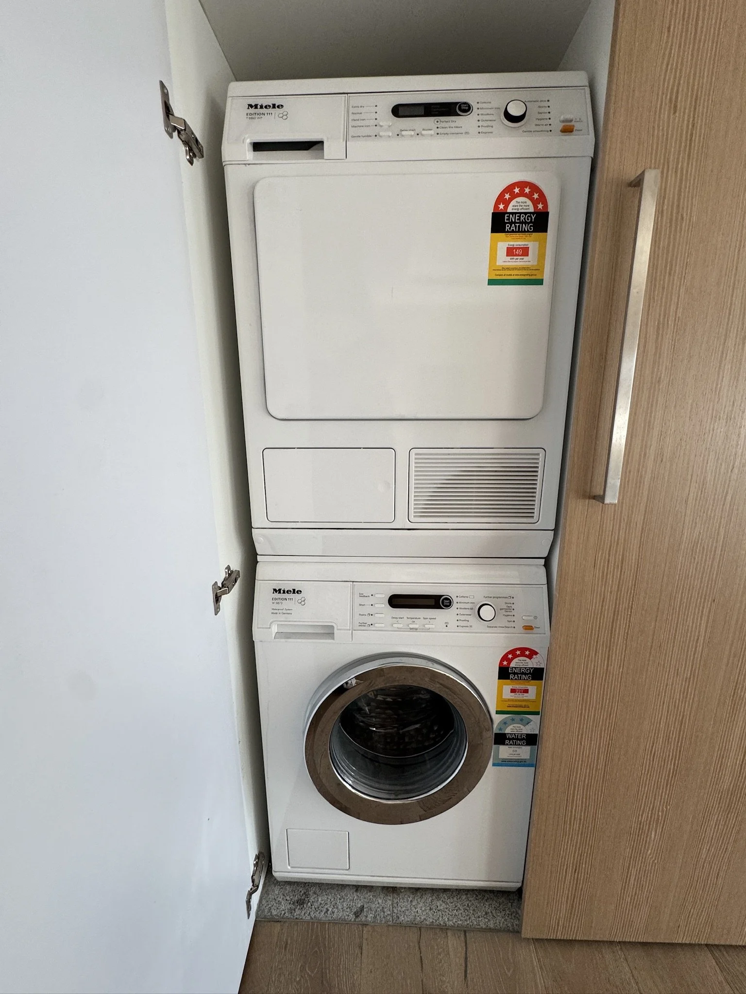 White front-loading washing machine and dryer stacked together in a laundry room, with a wooden cabinet to the right and a white wall to the left.