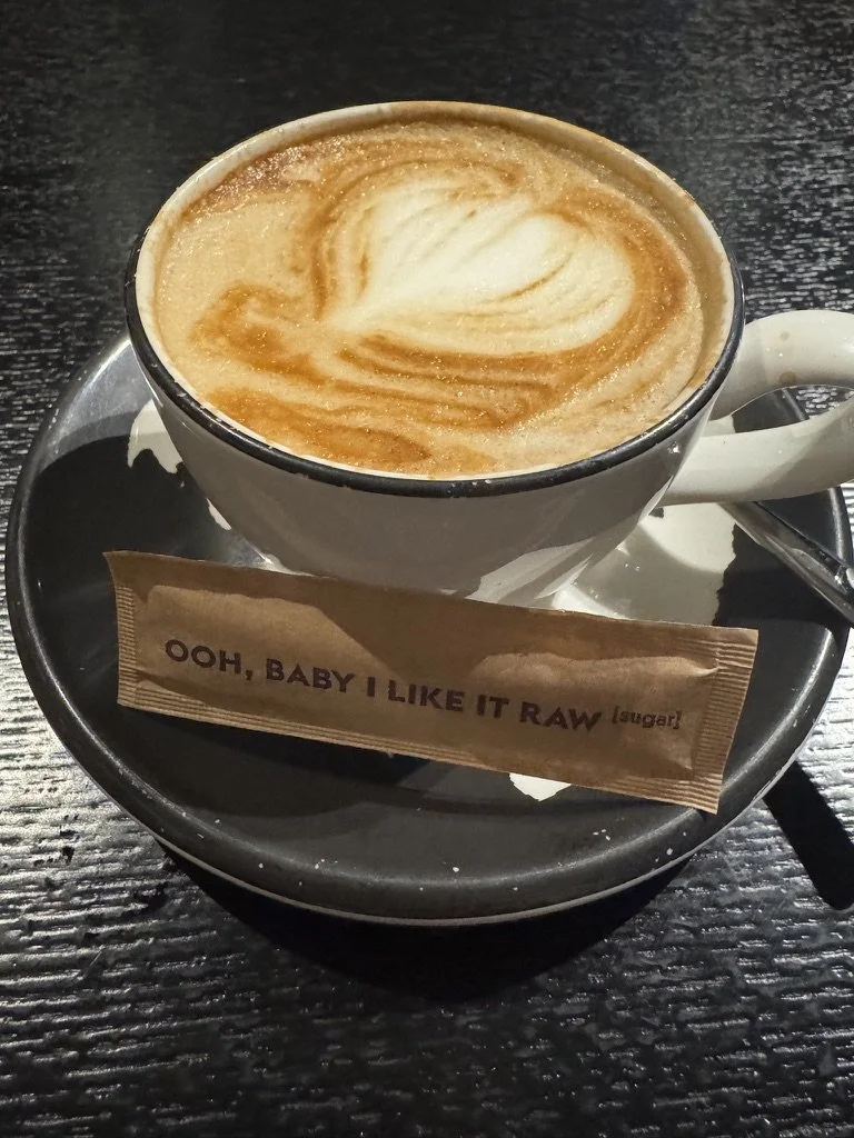 A cup of coffee with foam art on top served on a black saucer, with a sugar packet labeled 'Ooh, baby I like it raw' placed on the saucer.