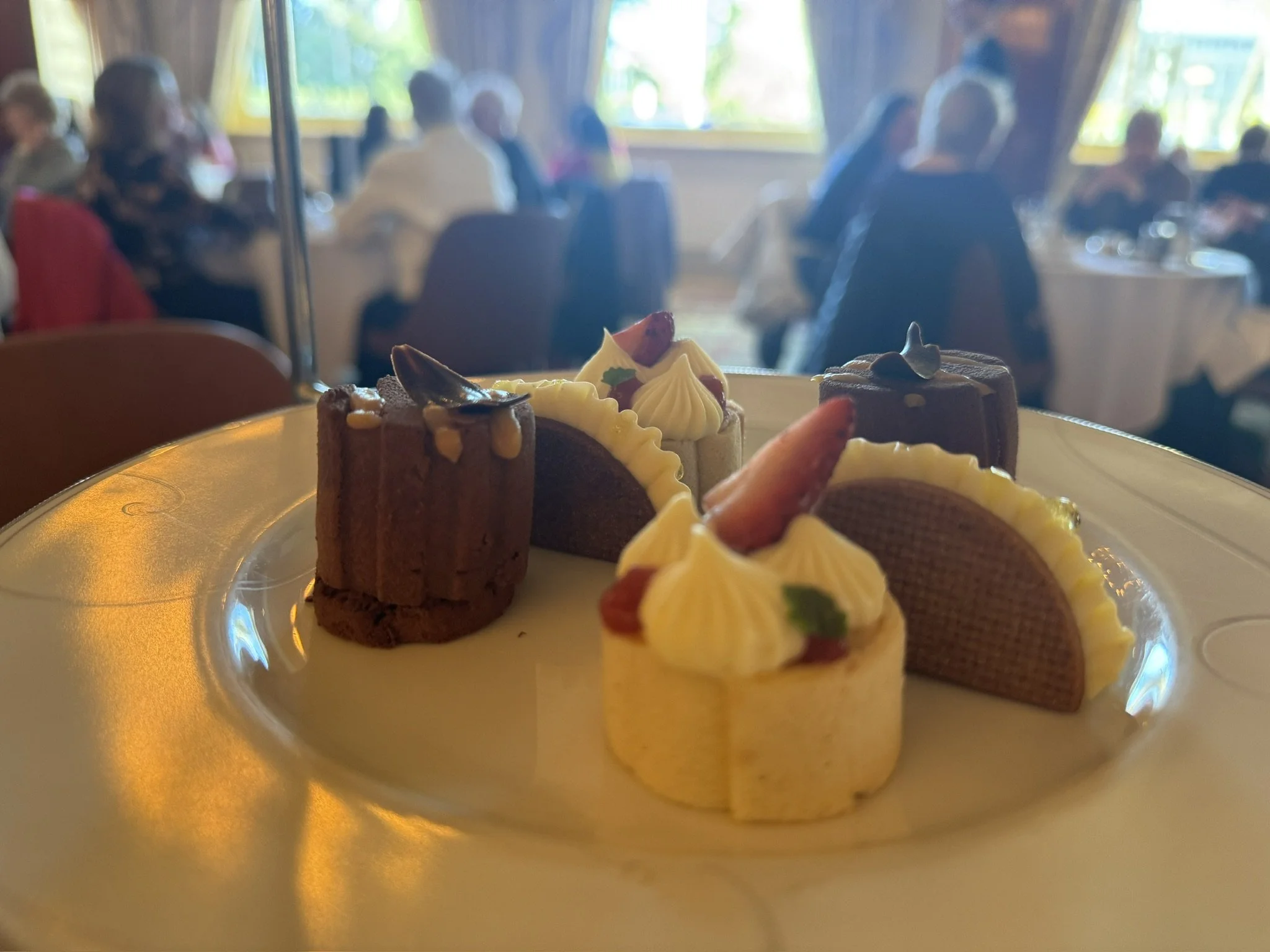 A plate of assorted desserts including cakes and pastries with cream and fruit garnishes, in focus in a brightly lit restaurant setting with blurred diners in the background.