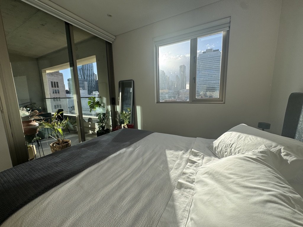 Sunlit bedroom with white bedding and large window showing city skyscrapers, with plants and a mirror near balcony.