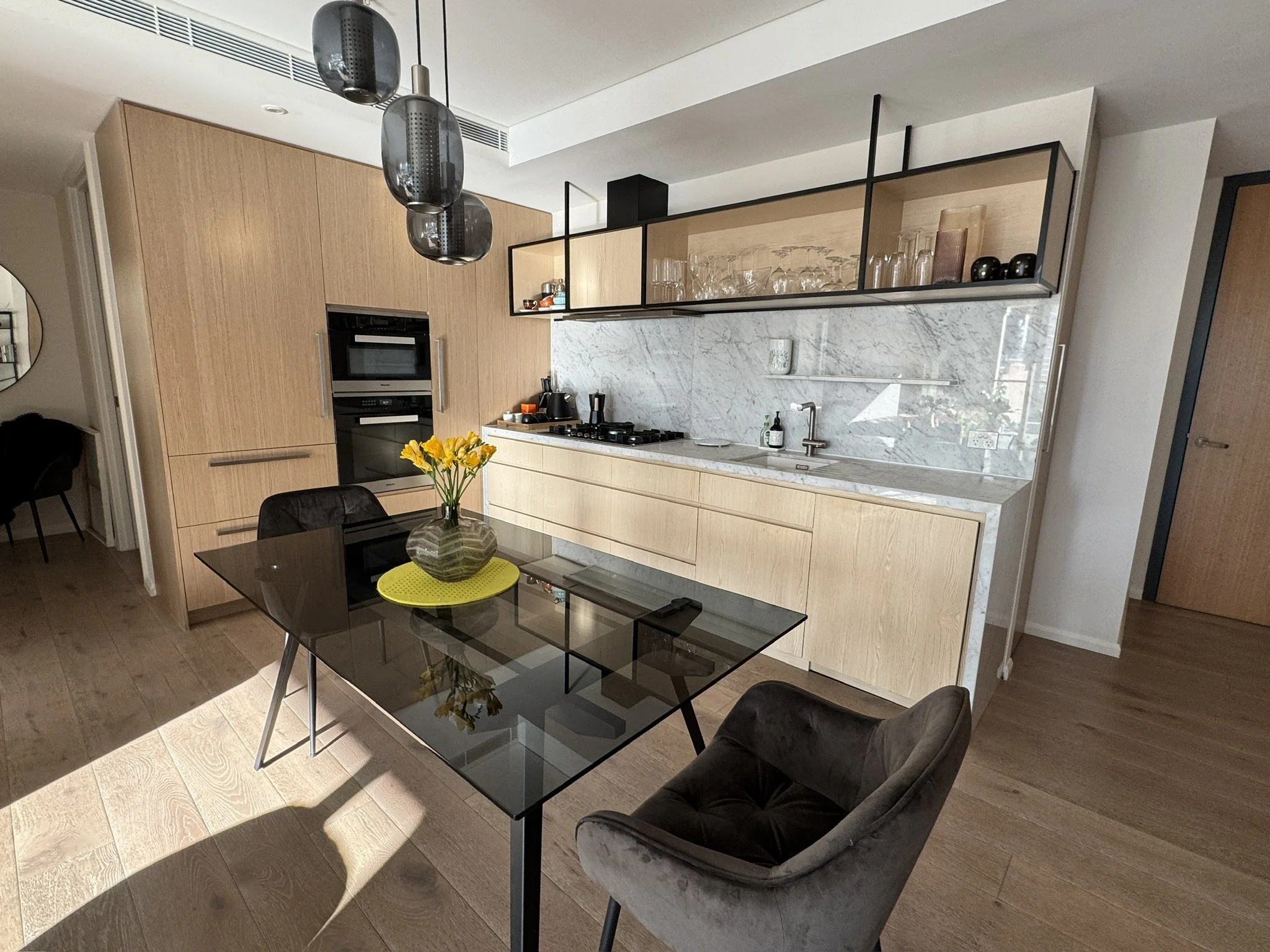 Modern kitchen with wooden cabinetry, marble backsplash, hanging pendant lights, glassware display, and a dining table with a vase of yellow flowers.