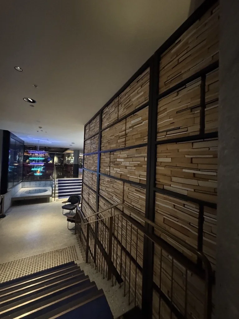Inside a modern building with staircase, a decorative wooden wall, and a seating area near a glass wall showing city lights at night.