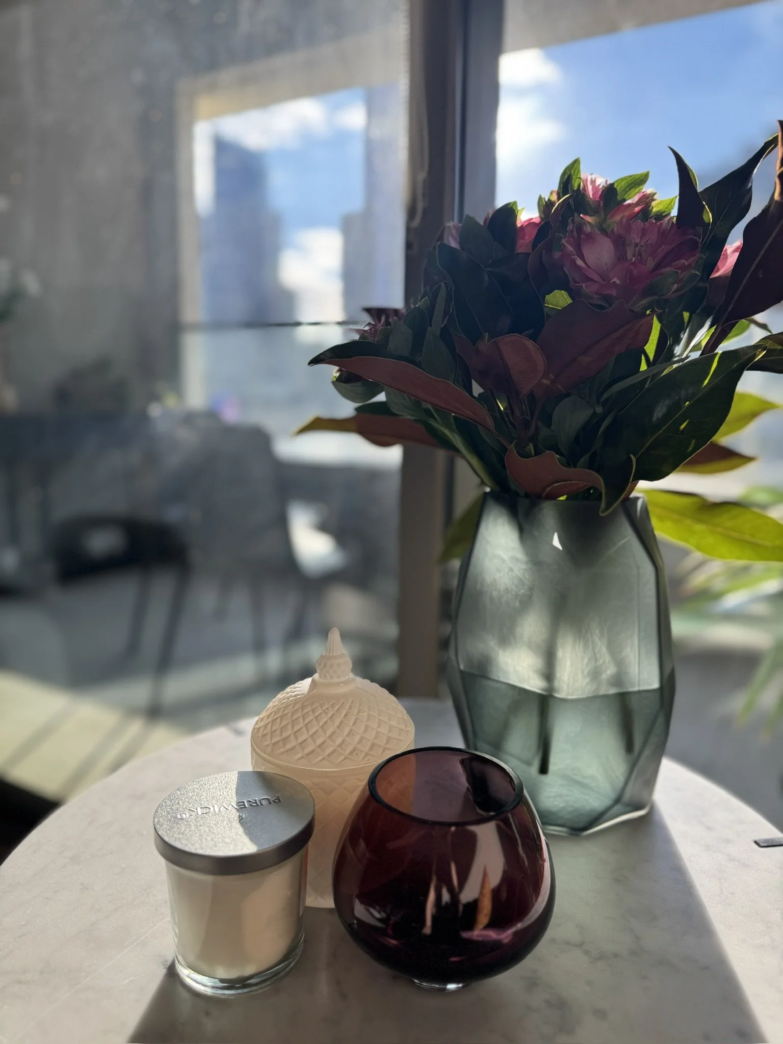 A vase with pink flowers, a small ice cream container, a red candle holder, a silver container labeled "Pure Milk," a glass container, and a round white object on a white table with sunlight coming through a window in the background.