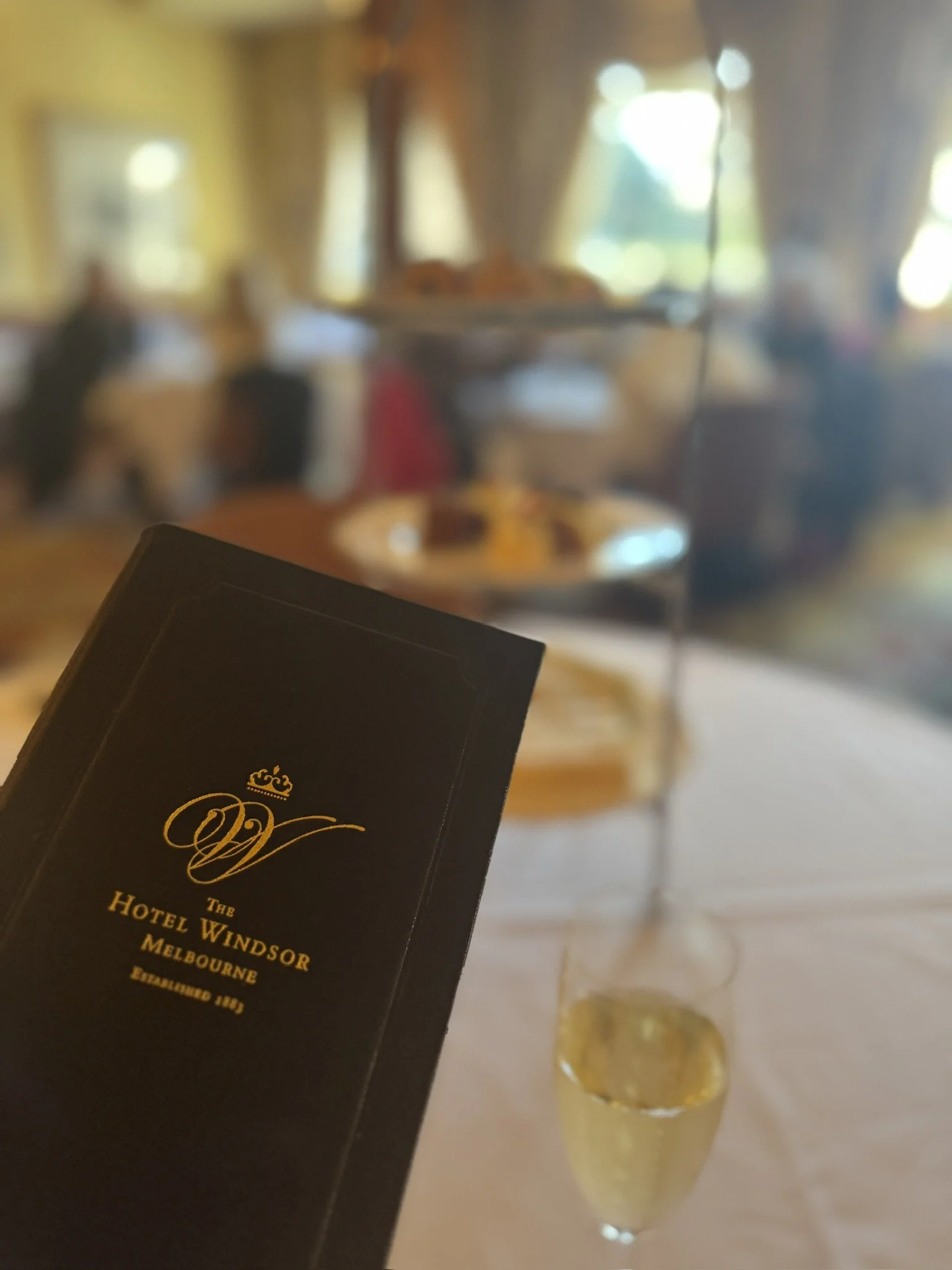 A black hotel room key card from the Windsor Hotel in Melbourne, with gold text and a crown logo, held near a champagne flute on a table with a blurred background of a well-lit room.