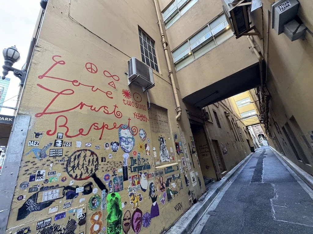Narrow alleyway with a mural on the left wall that reads "Love, Hope, Respect" in red cursive letters, decorated with various stickers, posters, and graffiti art. The alley is lined with buildings and leads to a brighter area ahead.
