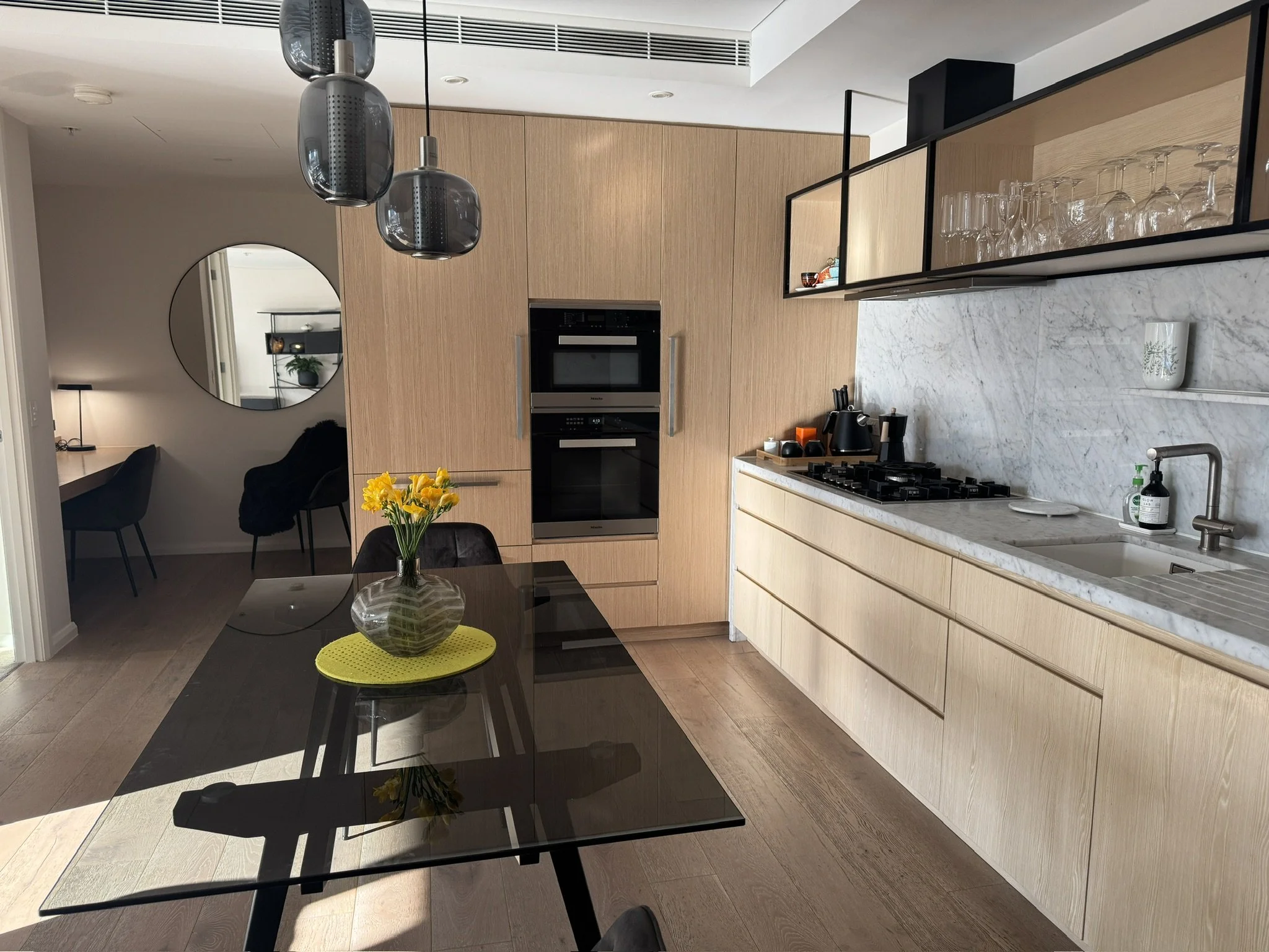 Modern kitchen with wooden cabinets, a marble countertop, glassware display, black dining table with yellow flowers, and pendant lights.