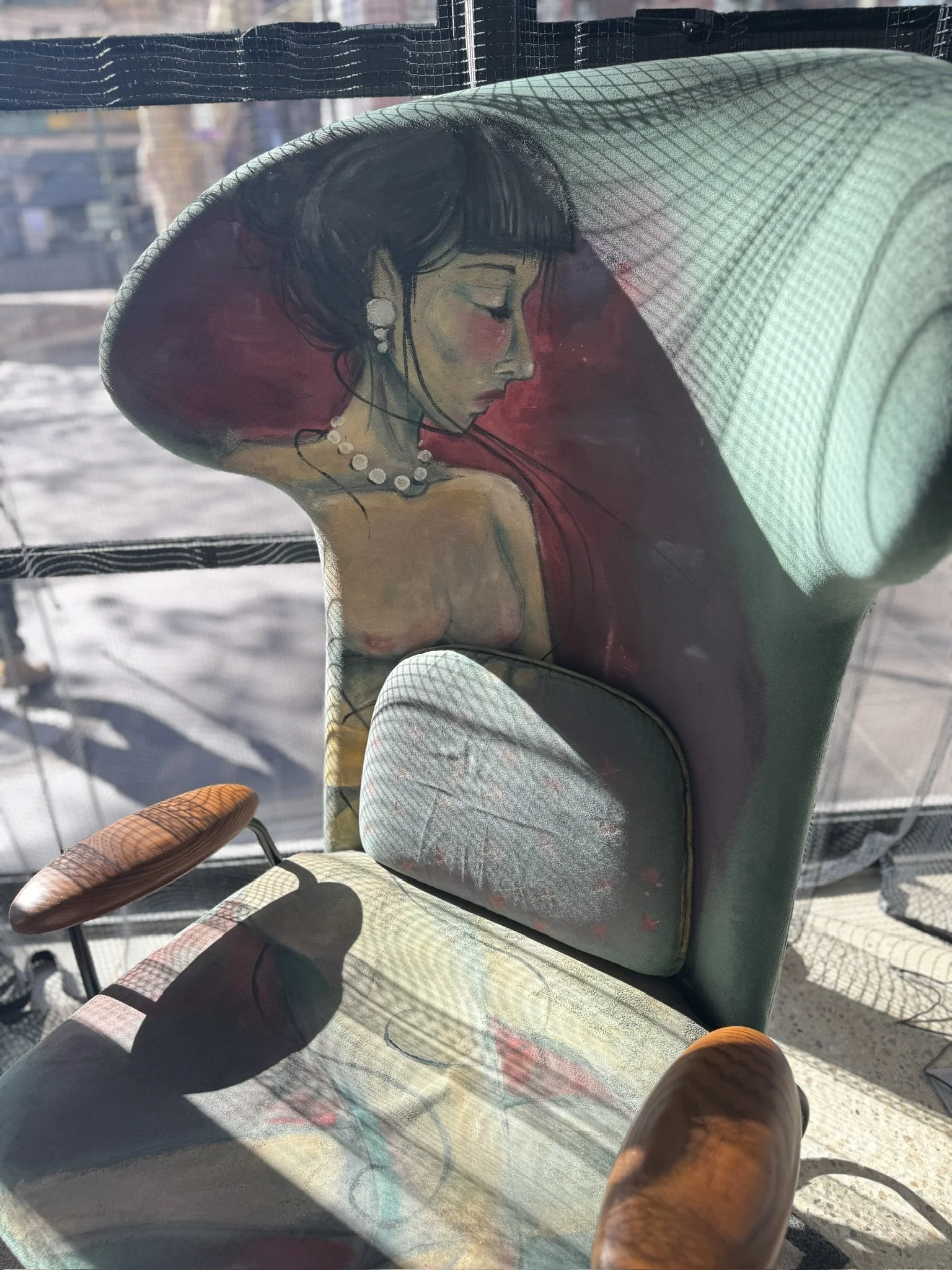 Painted illustration of a woman with black hair, wearing pearl earrings and a pearl necklace, in profile view with eyes closed and blush on cheeks, on a furniture piece with a light-colored cushion and wooden armrest, with sunlight casting shadows