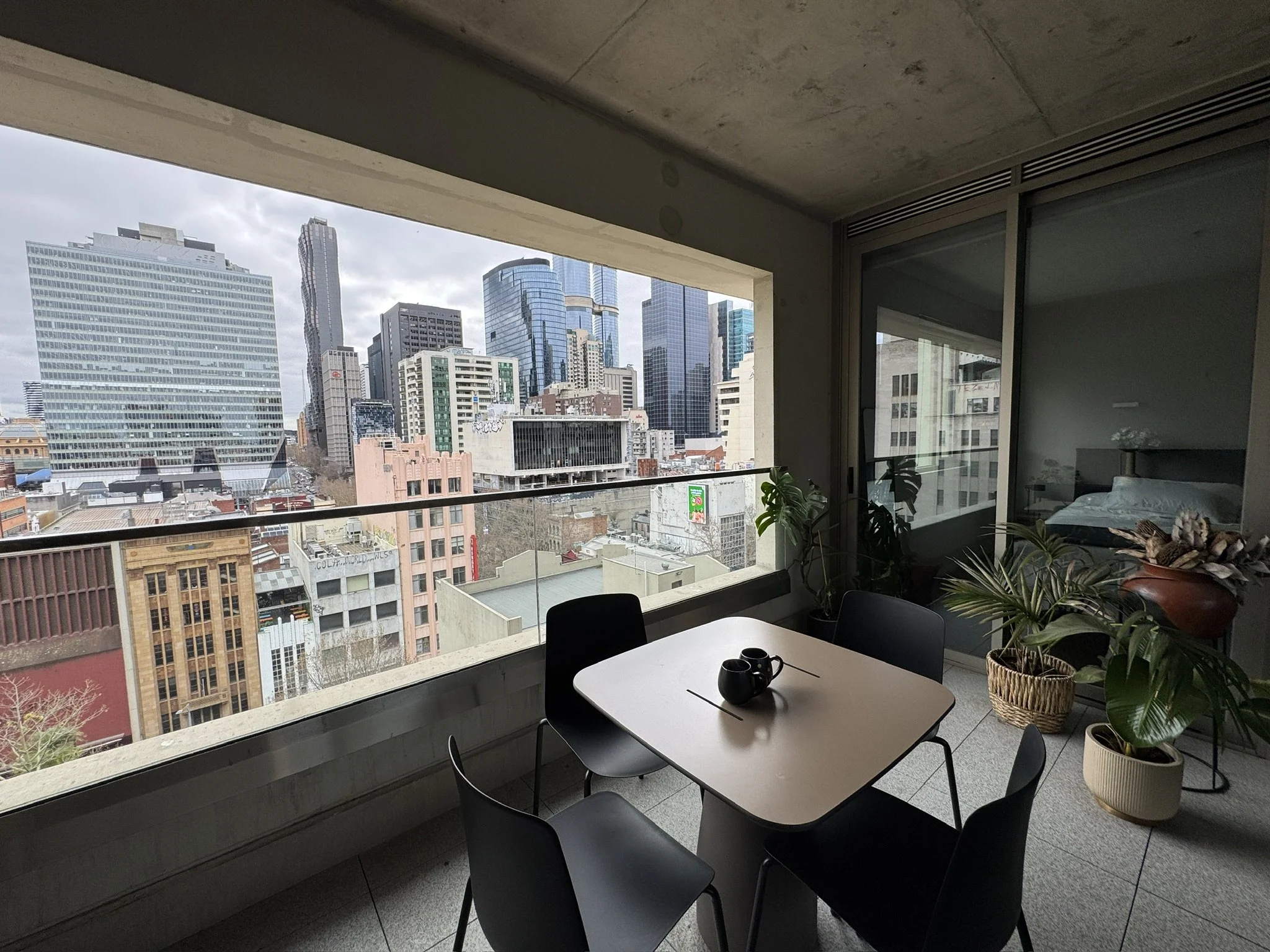 View from a balcony apartment showing a cityscape with modern high-rise buildings, some with glass facades, under a cloudy sky. Inside, there is a white table with black chairs, three black cups, and plants in a basket by the window.