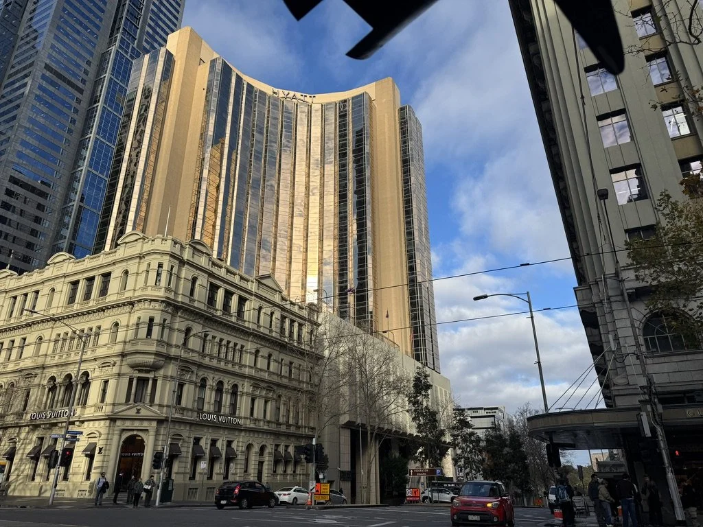 A view of tall modern and historic buildings in an urban city street with cars and pedestrians, under a partly cloudy sky.