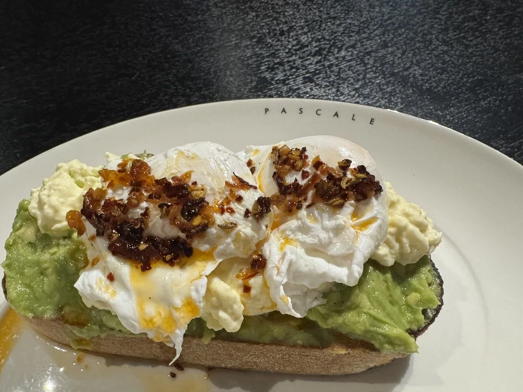 Open-faced breakfast sandwich with a toasted English muffin base topped with smashed avocado, poached eggs, and crispy bacon bits on a white plate with black background.