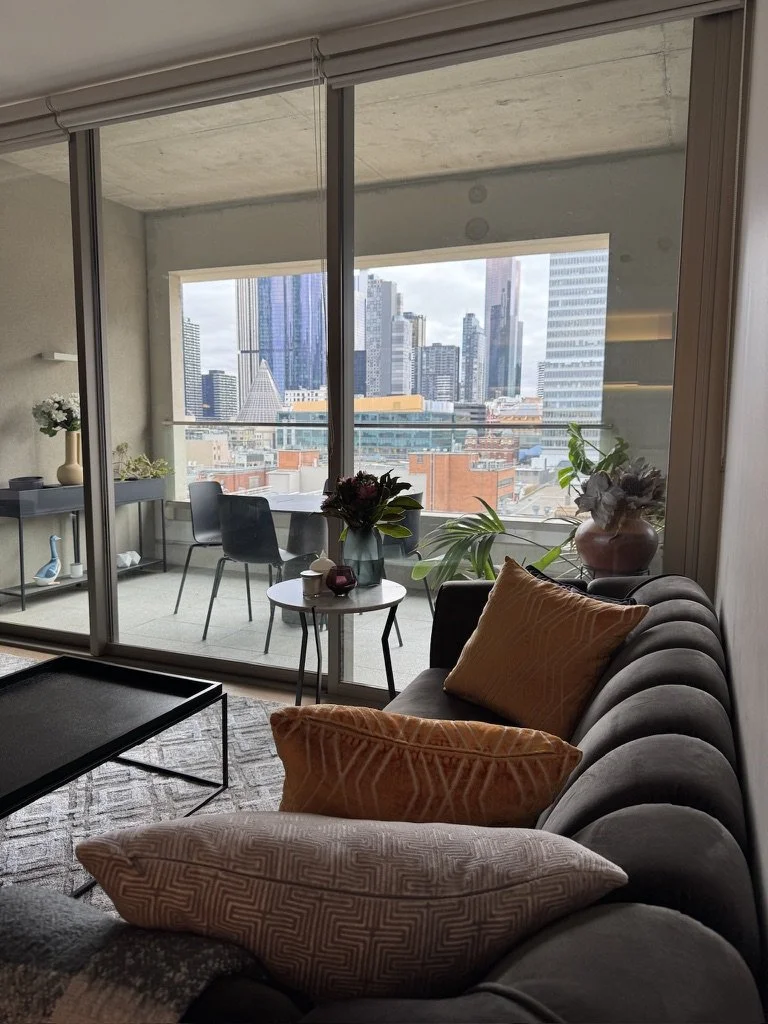 Living room with large glass windows overlooking a city skyline, decorated with potted plants, a gray sofa with orange and patterned throw pillows, a small side table with flowers, and a balcony with chairs and a table.