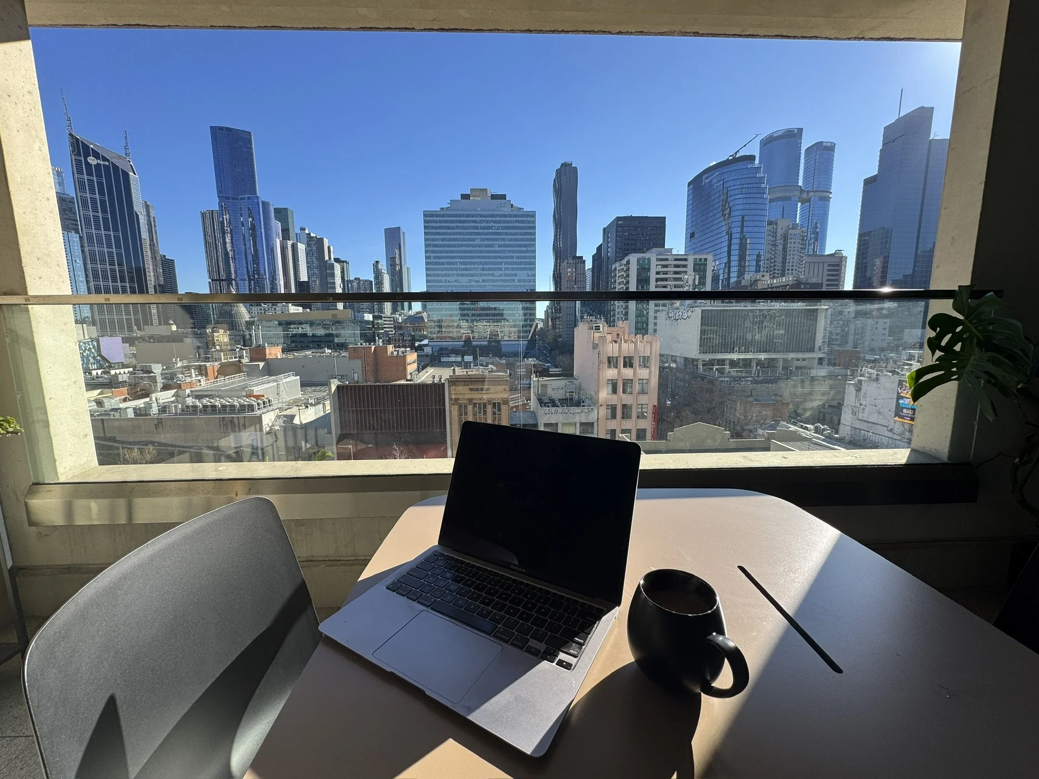 View of a city skyline with tall modern skyscrapers seen through a large window from inside an office or cafe, with a laptop, a mug, and chairs seen in the foreground.