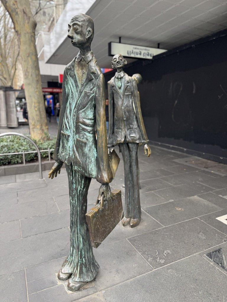 Bronze statues of two men in business suits standing on a city sidewalk, one holding a briefcase, with trees and buildings in the background.