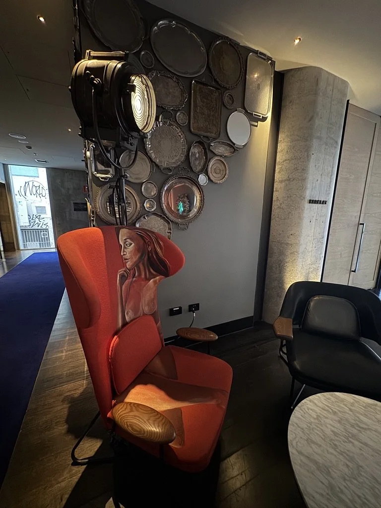 A red armchair with a painted portrait of a woman on its backrest, situated in a modern indoor space near a concrete wall and a collection of decorative plates and mirrors hanging on the wall.