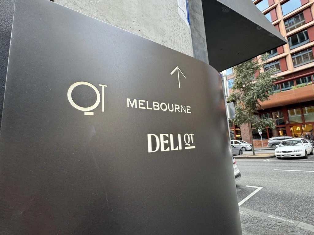 Urban street scene with a black sign indicating directions to Melbourne and a deli, with parked cars and modern buildings in the background.