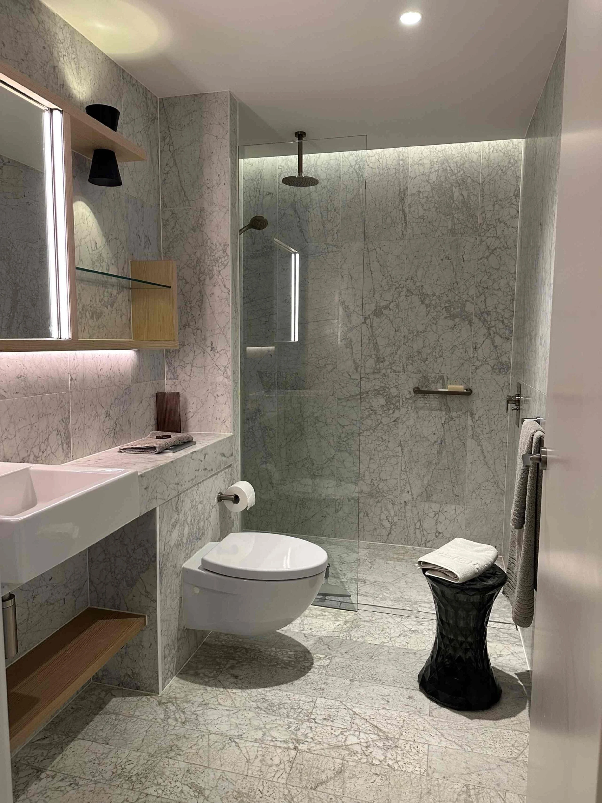 Modern bathroom with marble walls and floor, a toilet, a sink with a mirror, and a glass shower enclosure. There are towels hanging on a rail, a black trash bin, and a towel on the sink.