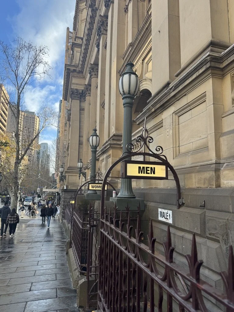 City sidewalk with street lamps and signs for men's and women's restrooms in front of a historic building.