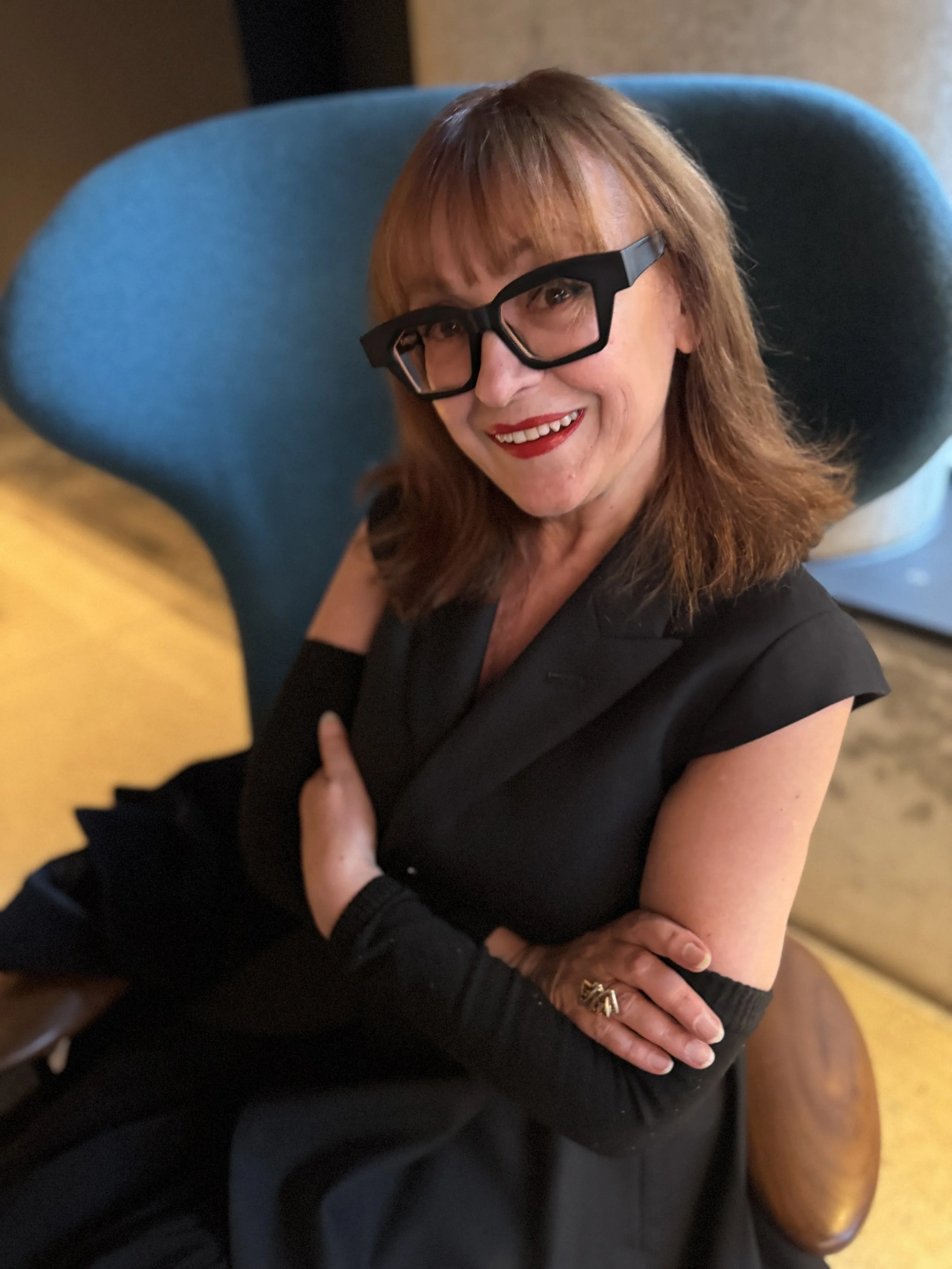 A woman with reddish-brown hair, wearing black glasses, red lipstick, and a black sleeveless top, smiling and sitting in a blue chair.