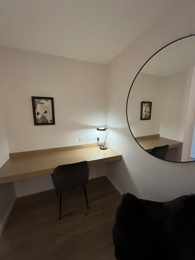 A minimalist desk area with a wooden desk mounted to the wall, a black chair, a small lamp, and a mirror on the wall reflecting the room.