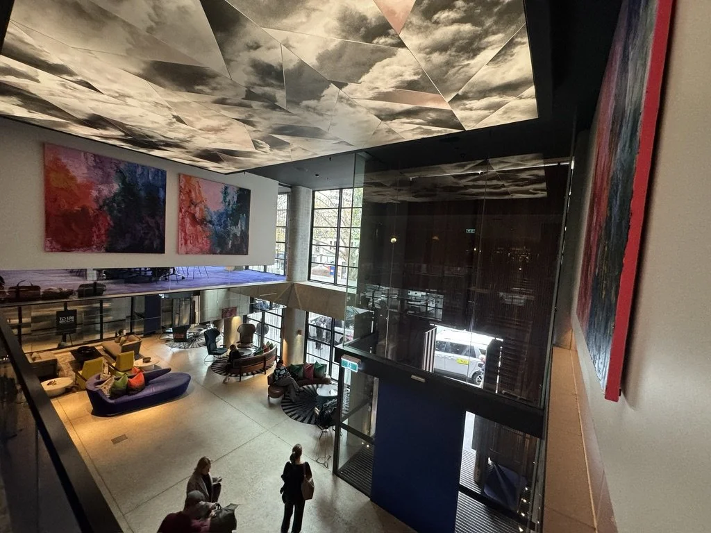 Modern hotel lobby with abstract paintings, floating ceiling art, and seating area with couches and chairs, some people walking and sitting.