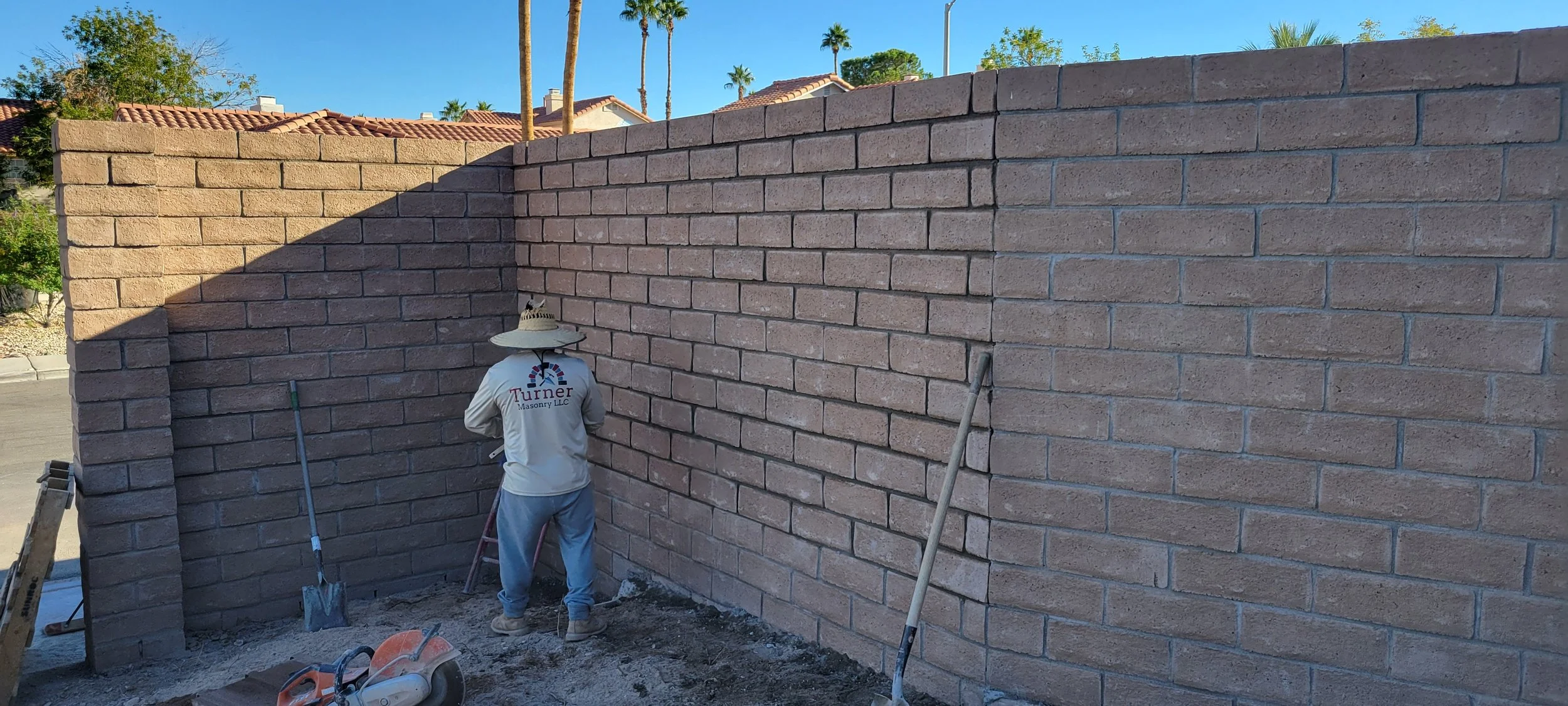 A construction worker building a brick wall in a backyard, with houses and palm trees in the background.