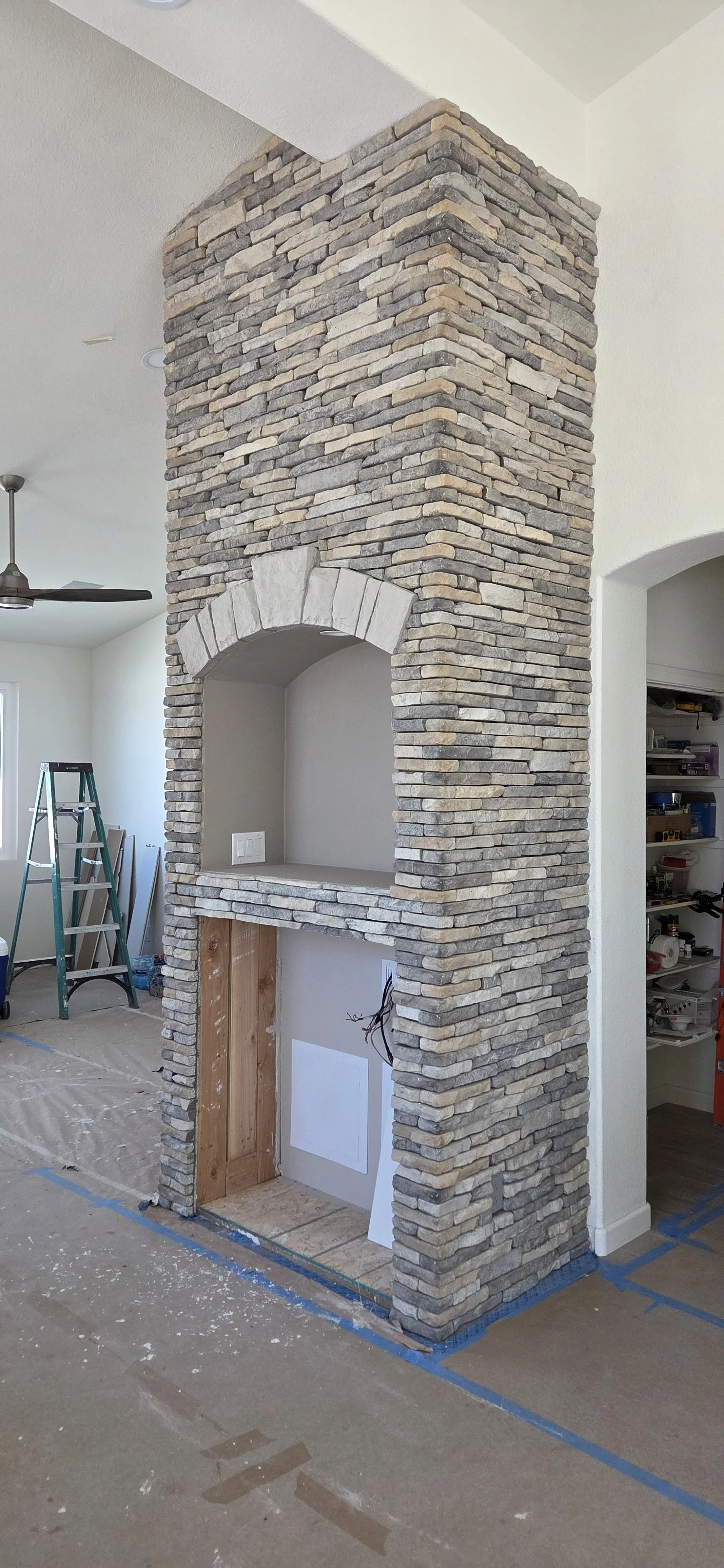 A partially constructed stone fireplace in a living space under renovation, with an arched alcove and a wooden base, surrounded by construction tools and supplies.