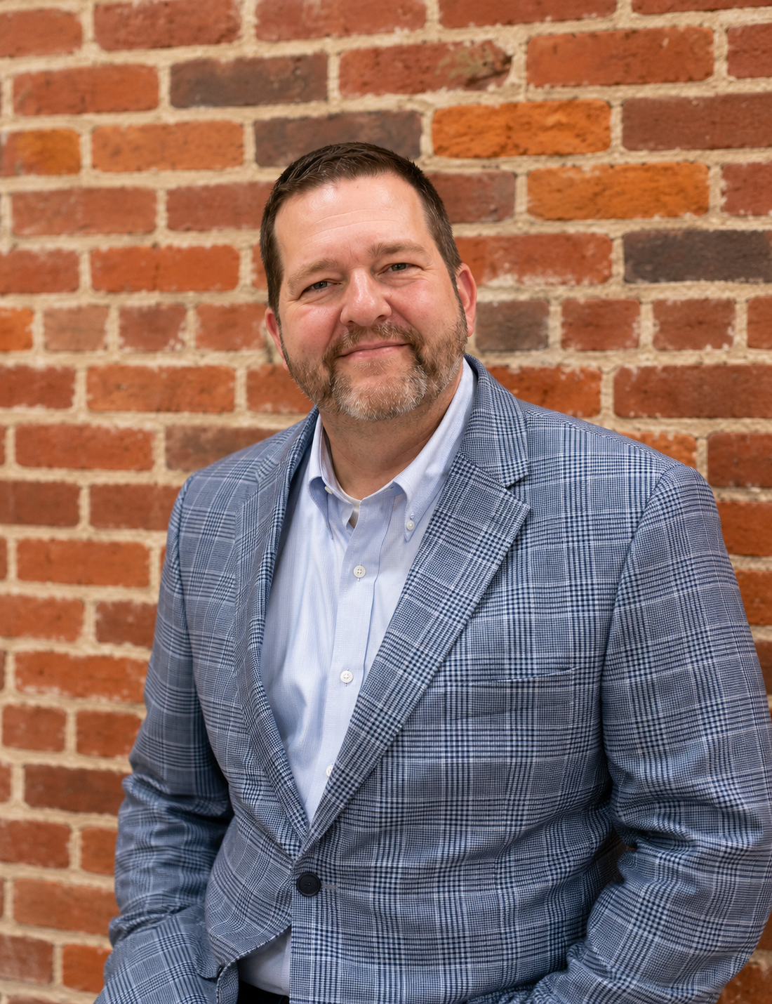 Man smiling in front of a brick wall, wearing a gray plaid blazer and a light blue shirt.