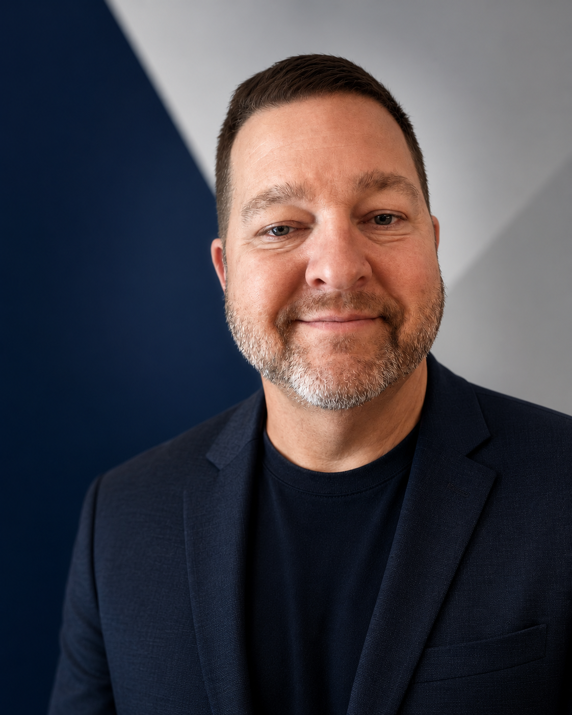 A middle-aged man with short brown hair and a beard, wearing a dark blazer and black shirt, smiling at the camera against a background with dark blue and light gray geometric patterns.