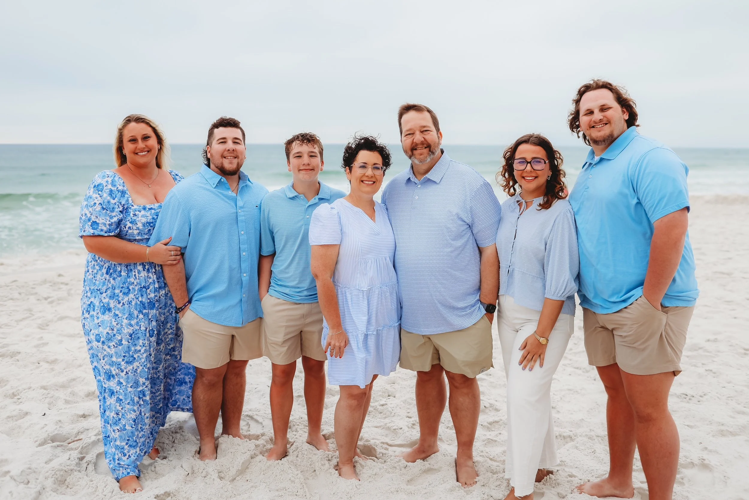 A group of eight people smiling at the beach, standing on the sand with the ocean in the background, dressed in casual summer clothing in shades of blue and white.