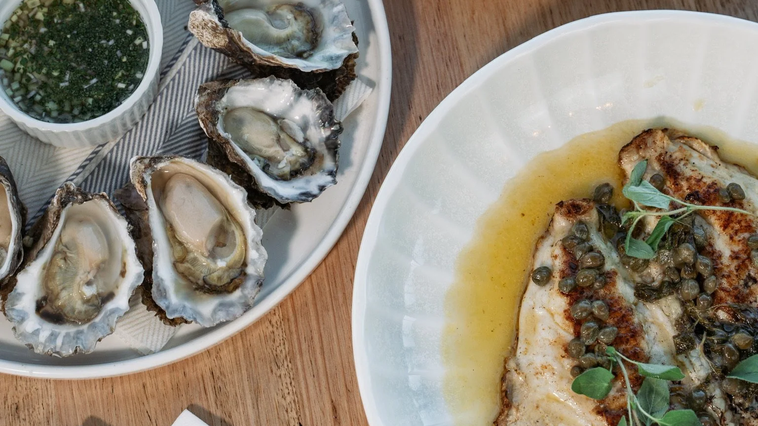 Close-up of a plate of cooked oysters topped with capers and fresh herbs, served with lemon butter sauce, alongside a dish of raw oysters with a cup of green sauce.