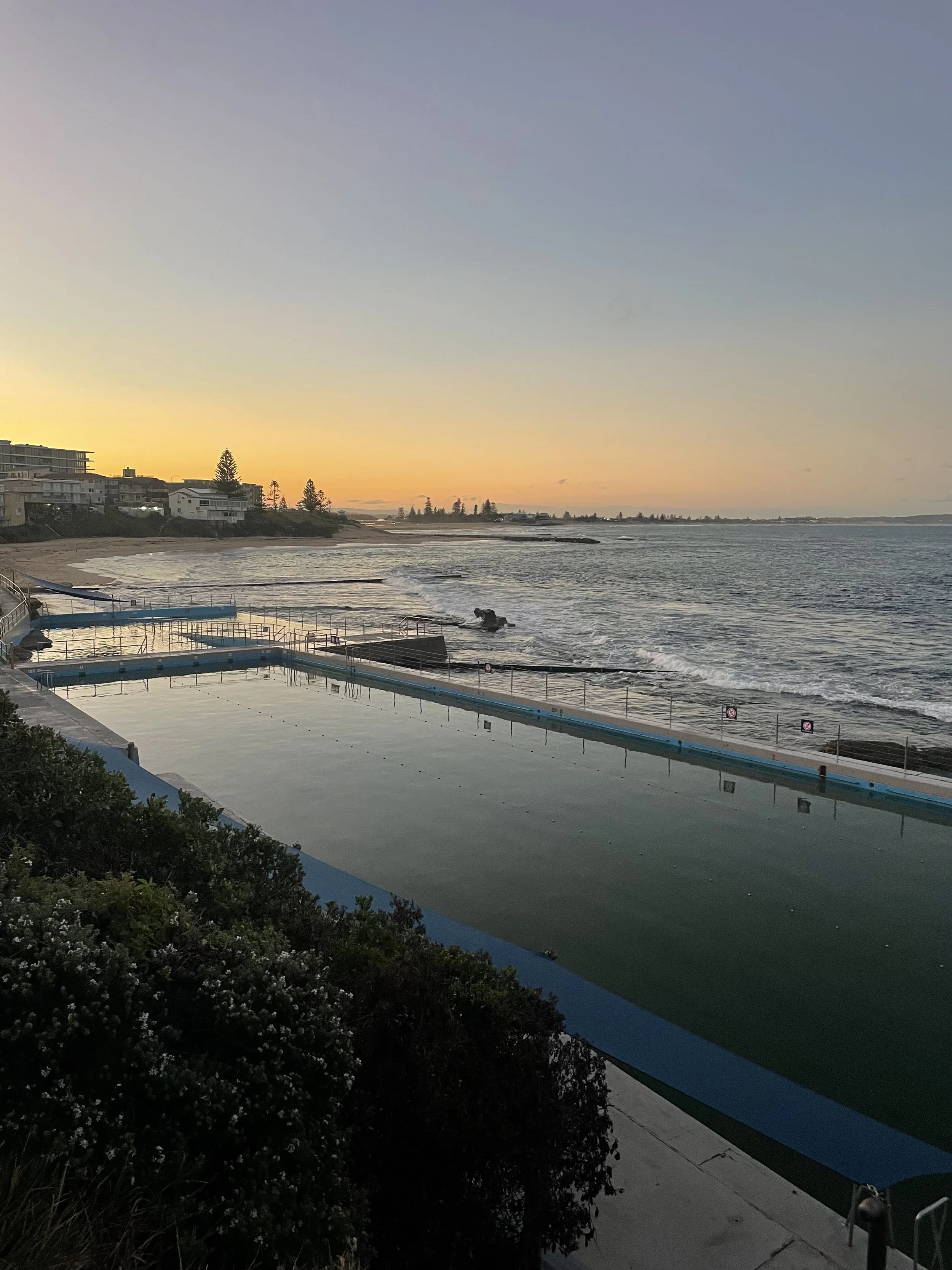 Sunset view over the ocean with a beach, residential buildings on the left, and an infinity pool in the foreground at dusk.