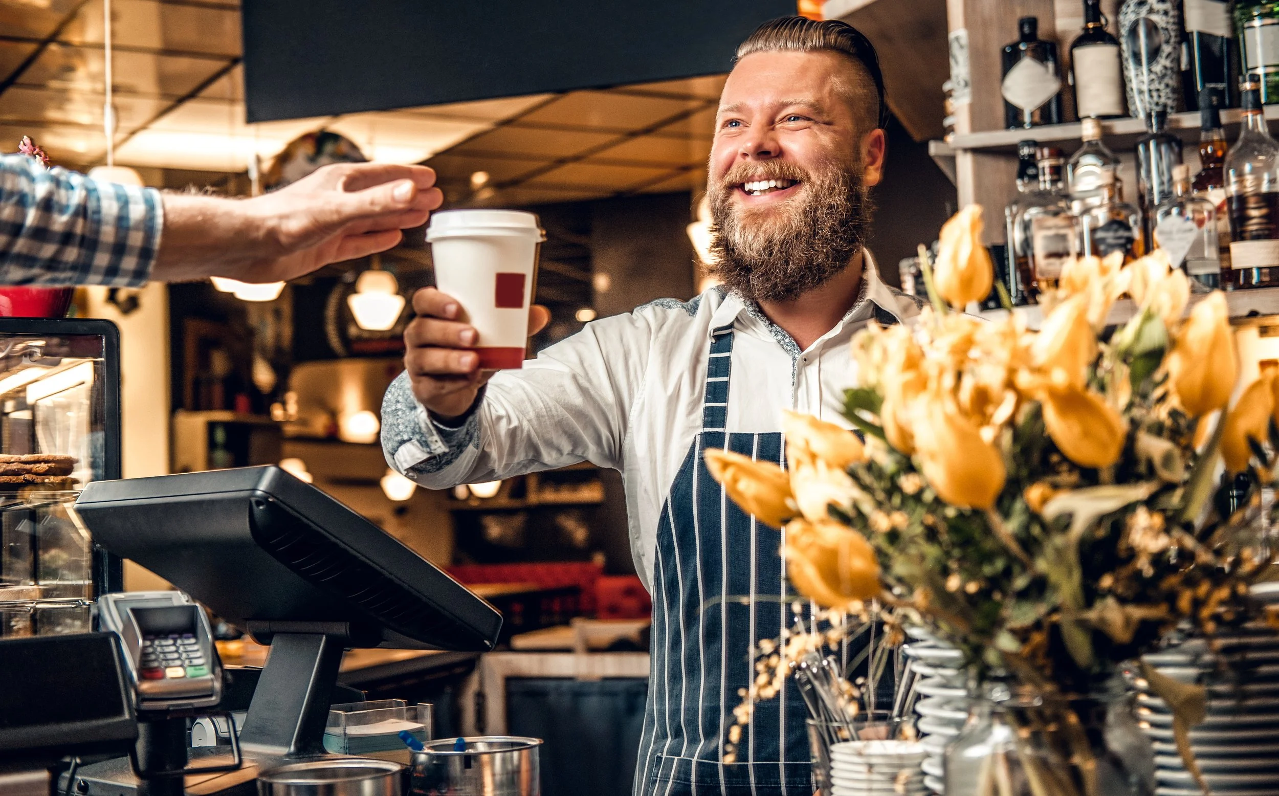 A smiling barista with a beard wearing a striped apron handing a coffee cup to a customer at a coffee shop counter with flowers in the foreground.