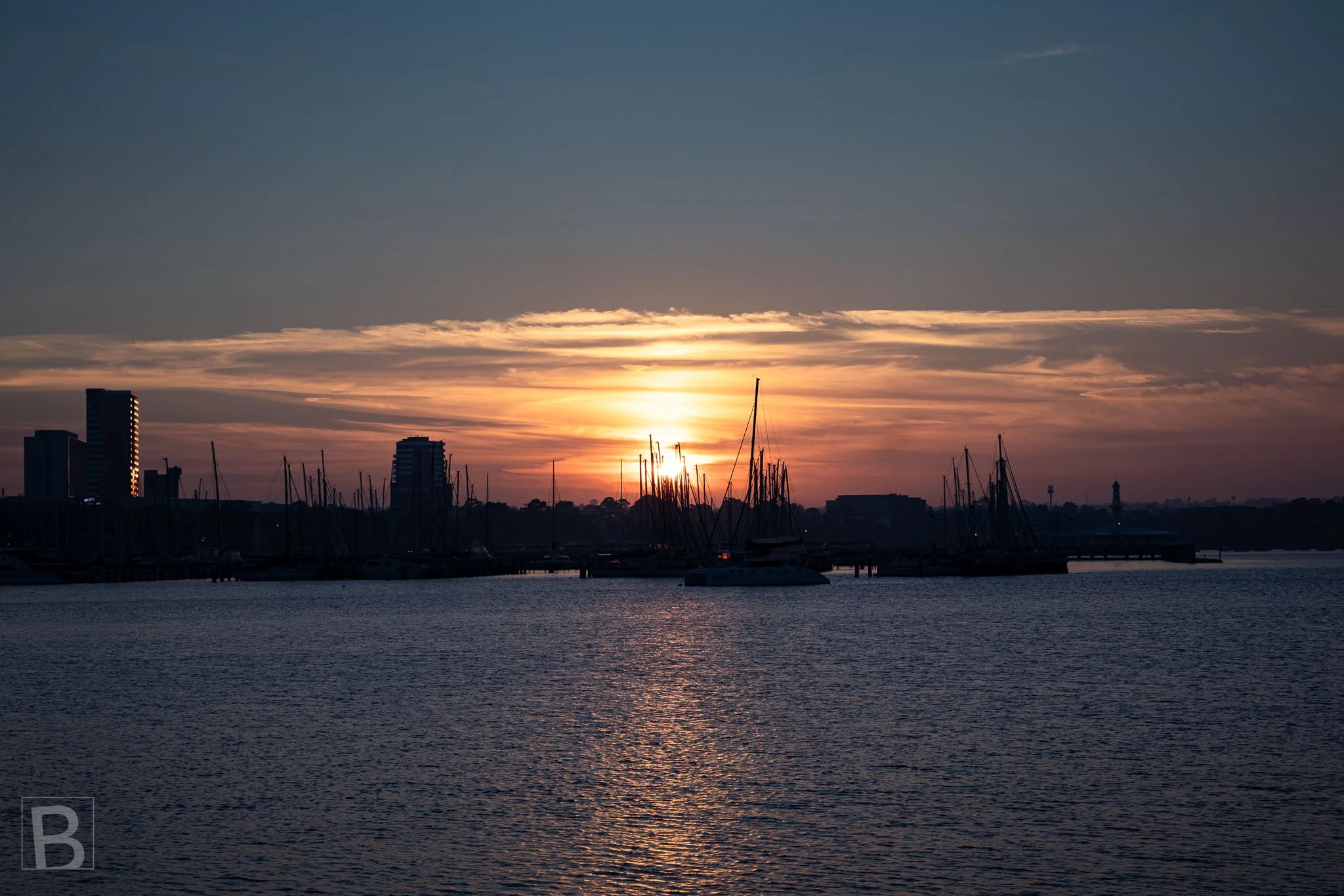 The sinking sun illuminates a harbour horizon, tracing warm light
between silhouetted masts and across the evening water.