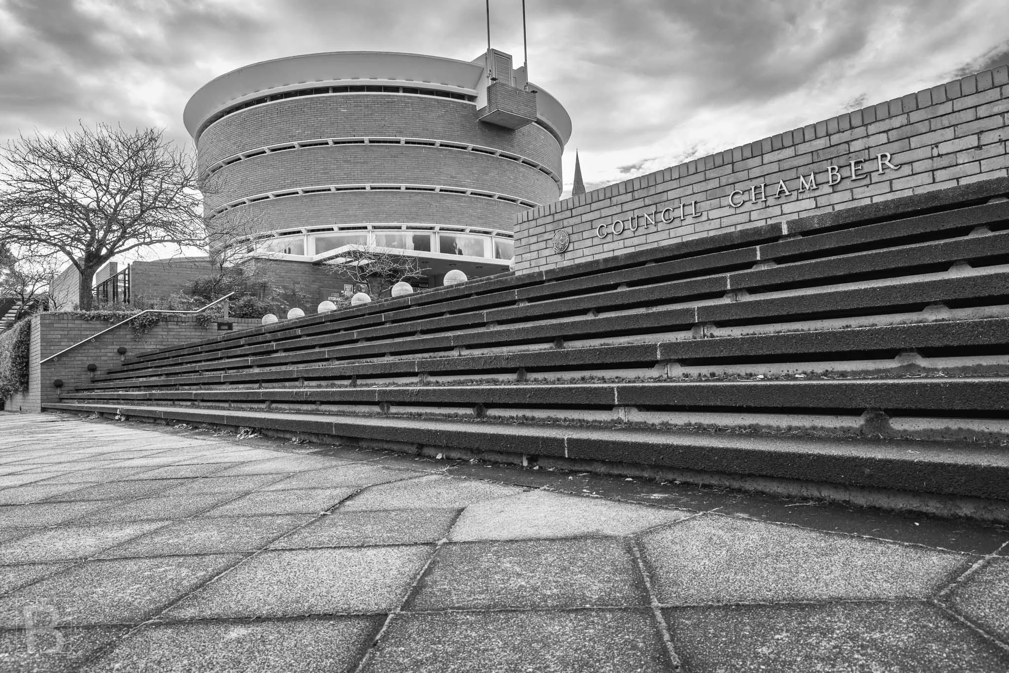 Black and white photograph of the Brighton Council Chamber in Melbourne, showing the curved brick civic building behind long stepped terraces and the Council Chamber wall.