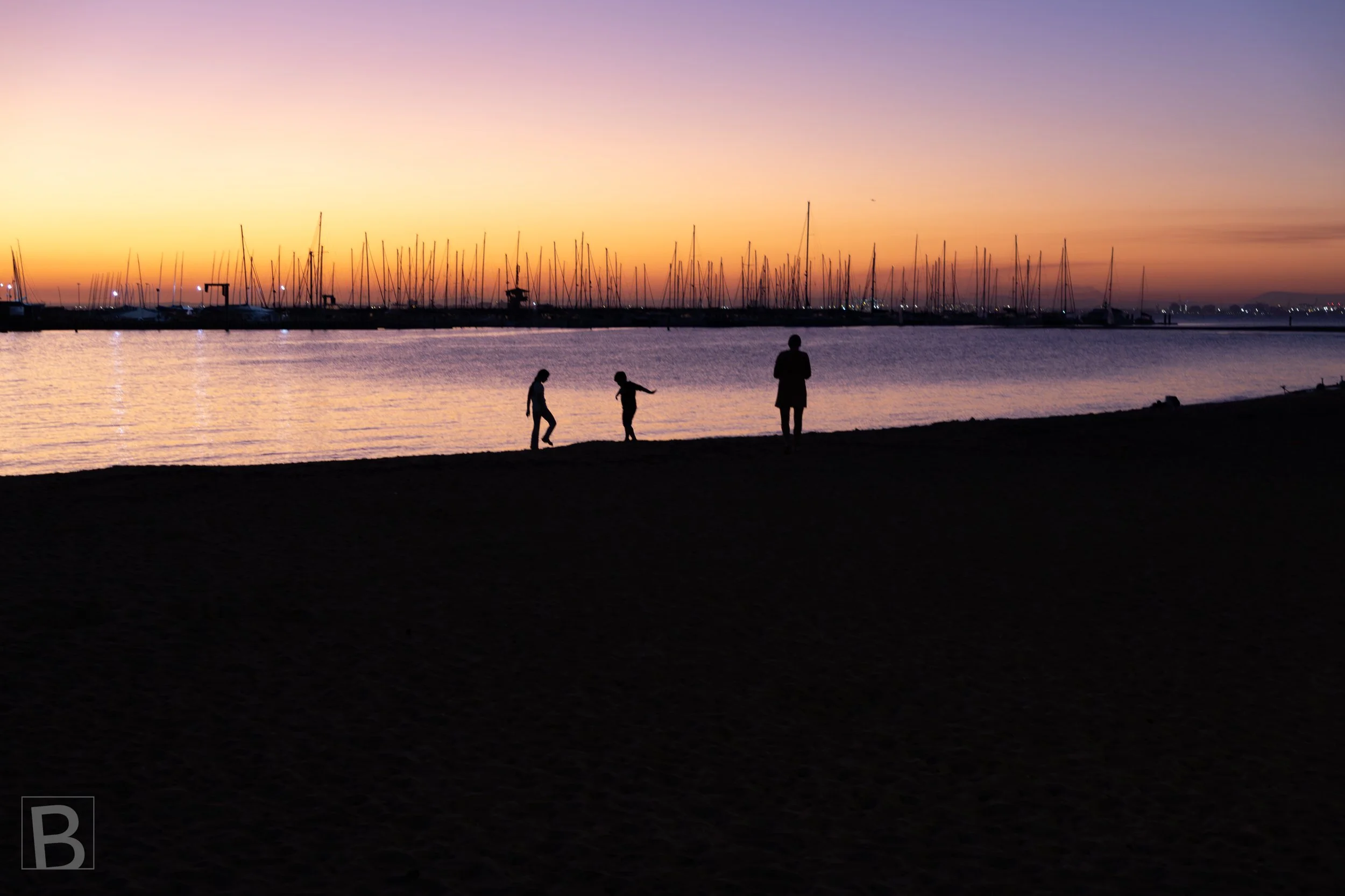 Small figures pause at the water's edge as pastel evening light fades
behind a forest of marina masts.
