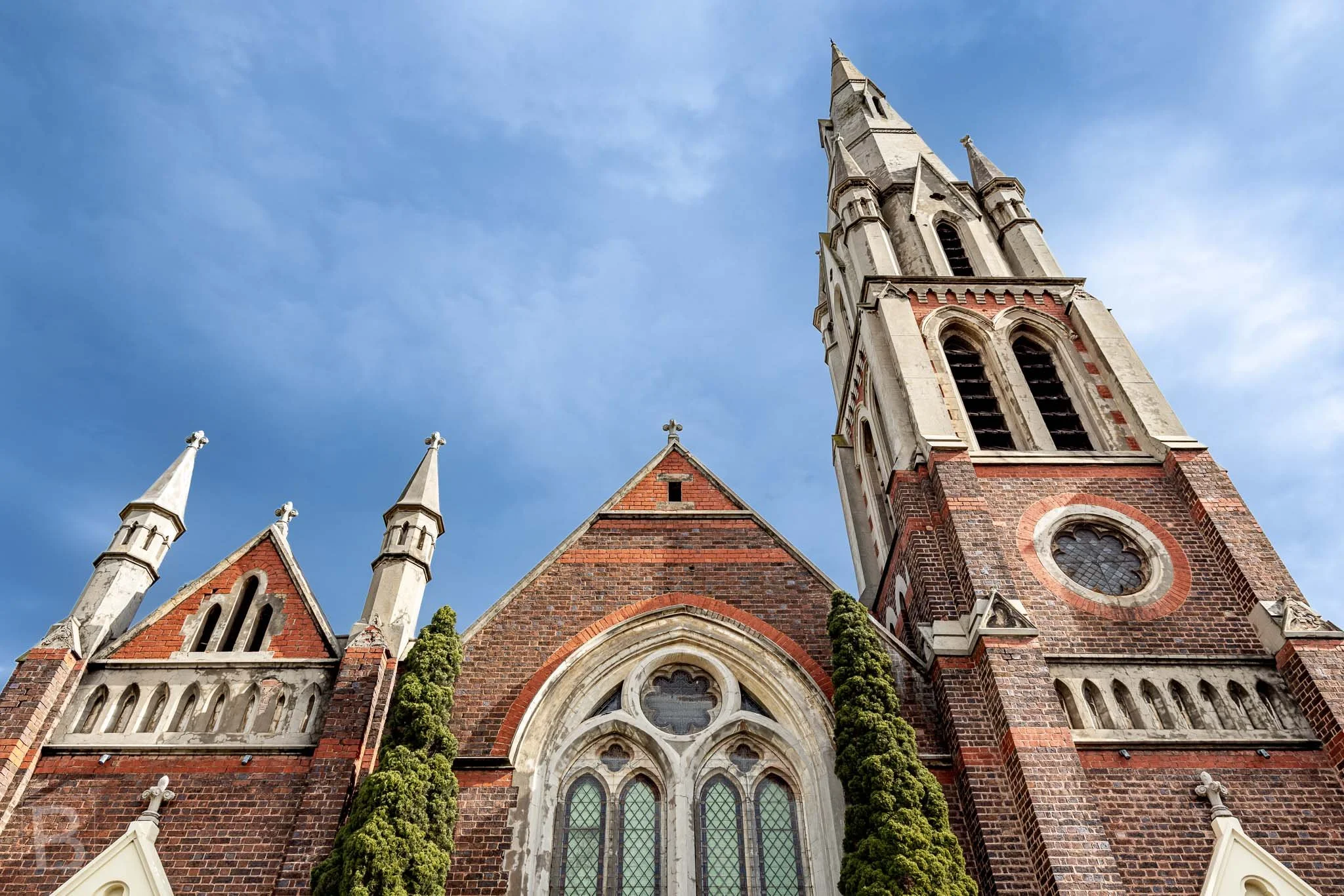 Upward view of a red-brick heritage church in Brighton, Melbourne, showing Gothic arches, decorative stonework and the tower.