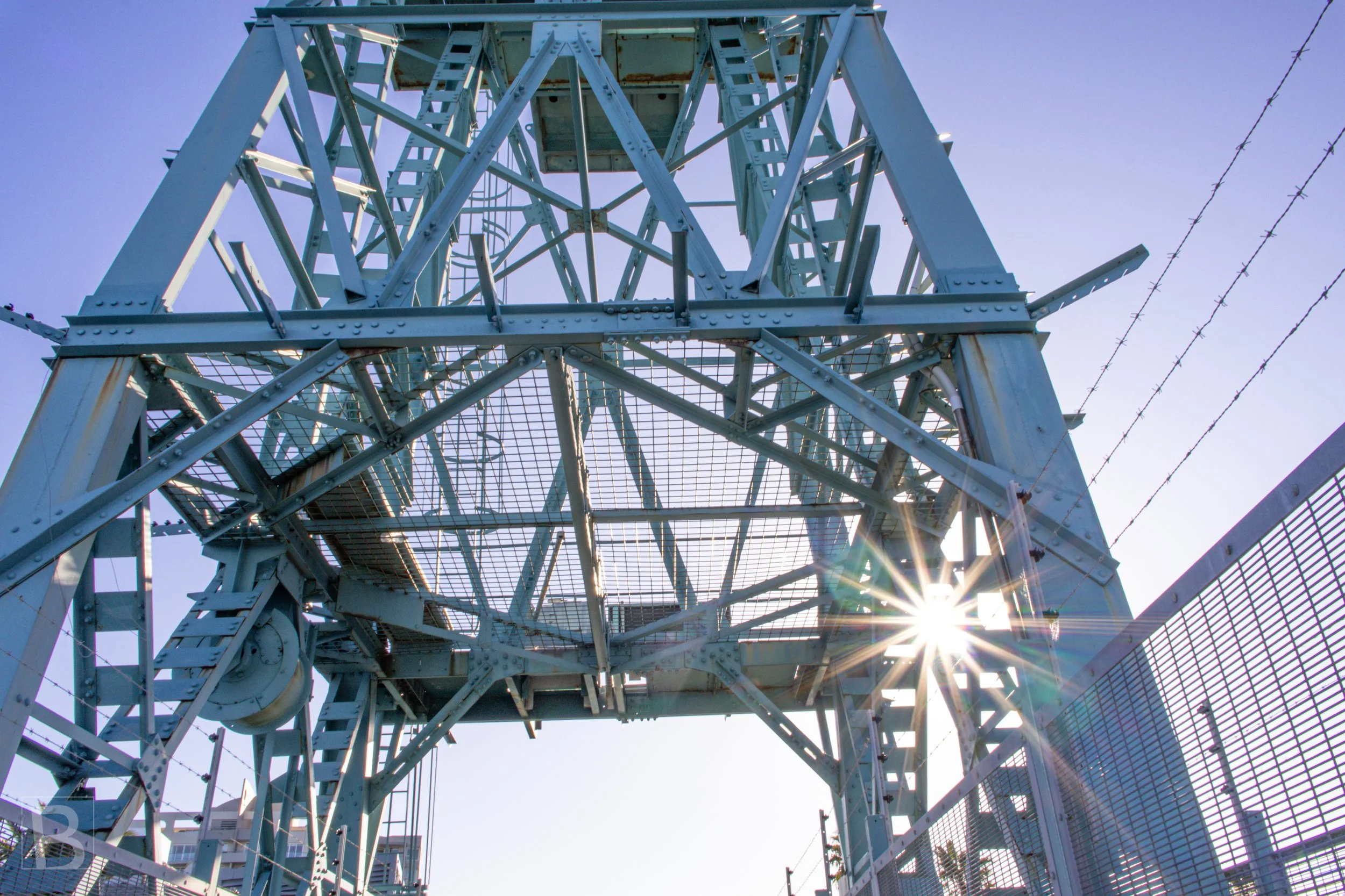 A sharp sunburst reveals the intricate lattice, platforms and cables of
a working crane from directly below.