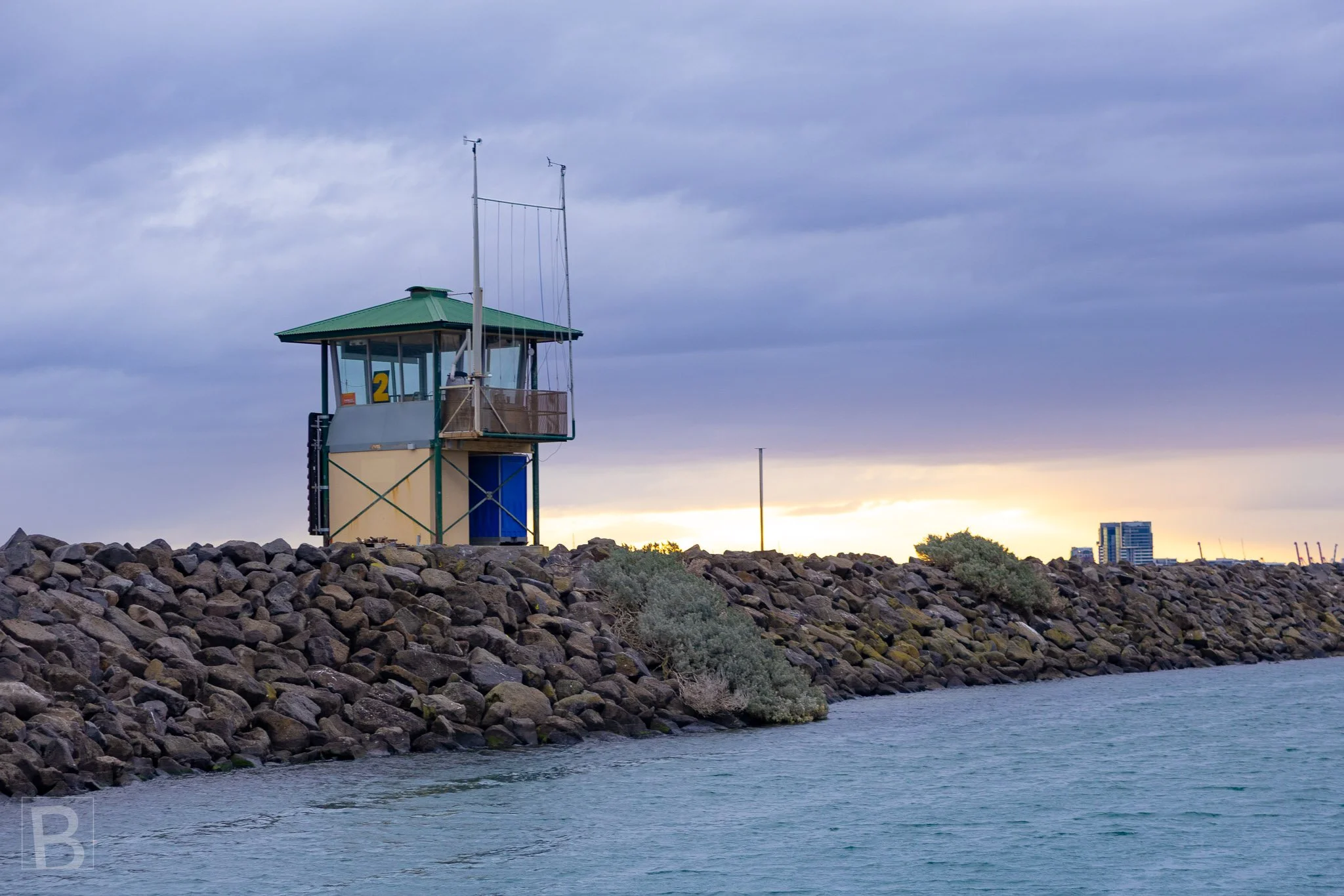 A compact harbour structure keeps watch from the breakwater, its
angular form set against rock, water and approaching weather.