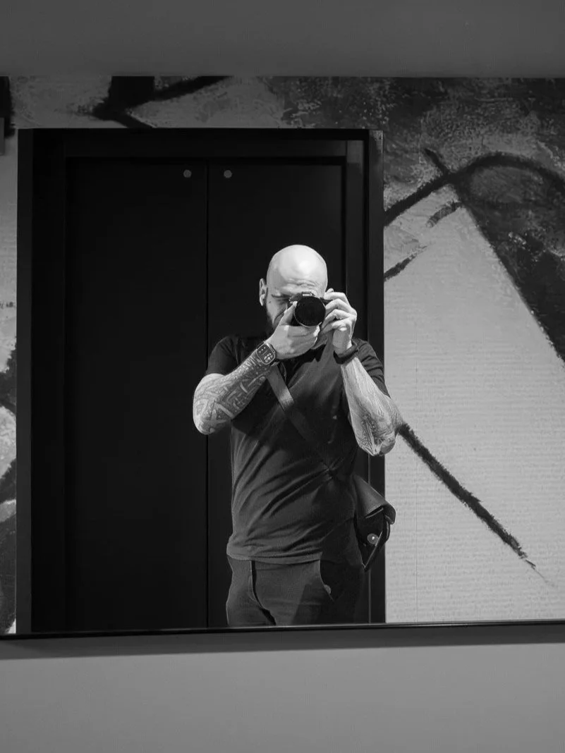 Byron taking a selfie with a camera in front of a mirror, tattoos on arms, wearing a black t-shirt, with an abstract mural on the wall behind him.