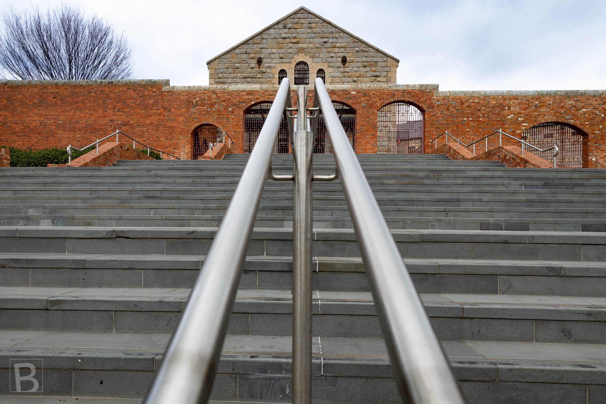 Symmetrical stairs and metal handrails lead upward towards the stone and red-brick architecture of Bendigo's former Sandhurst Gaol complex.