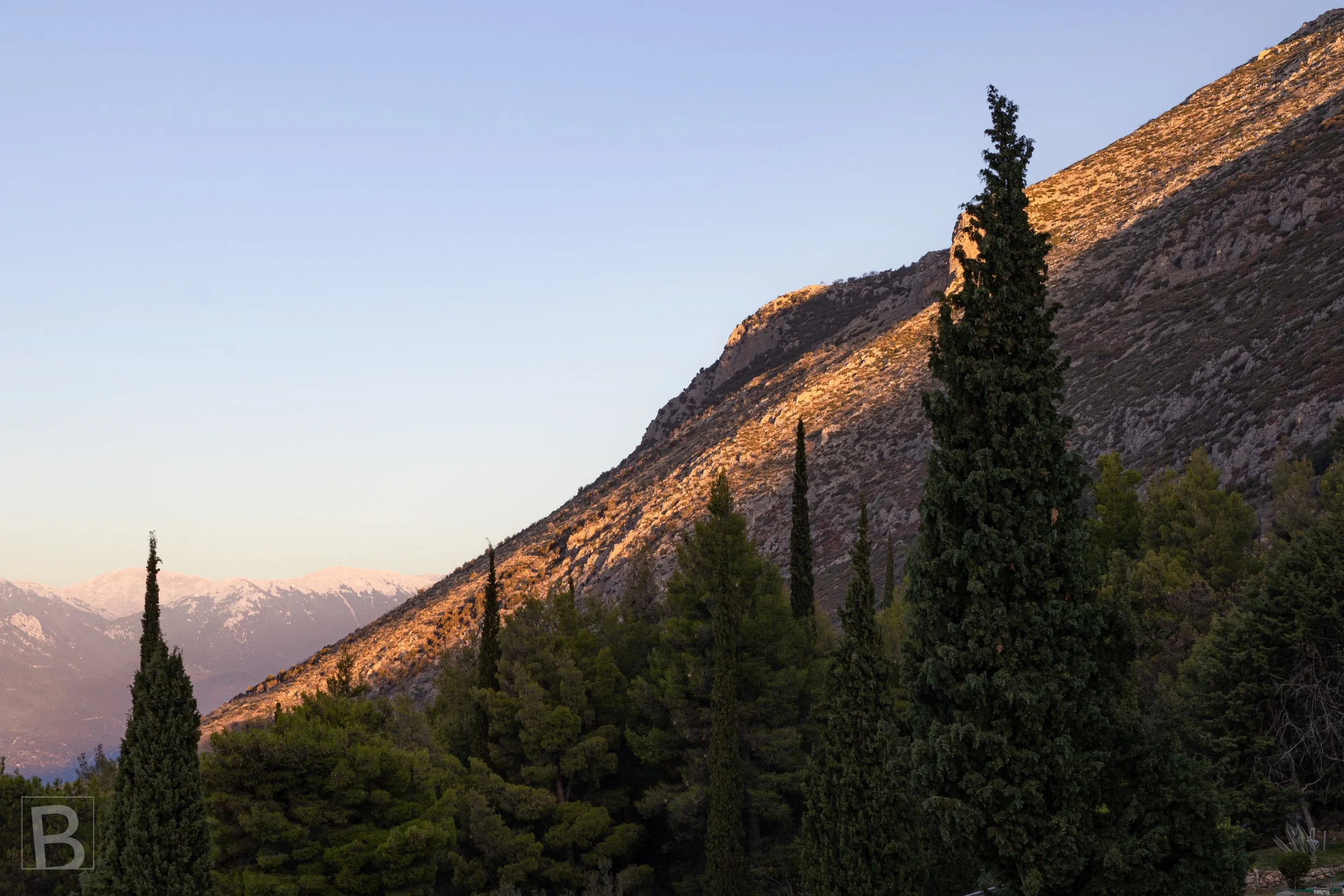 A steep mountain slope catches the final warm light of day, rising
above cypress trees and softly receding ranges.Mount Parnassus catches the final warm light of day, rising above dark cypress trees and softly receding mountain ranges.