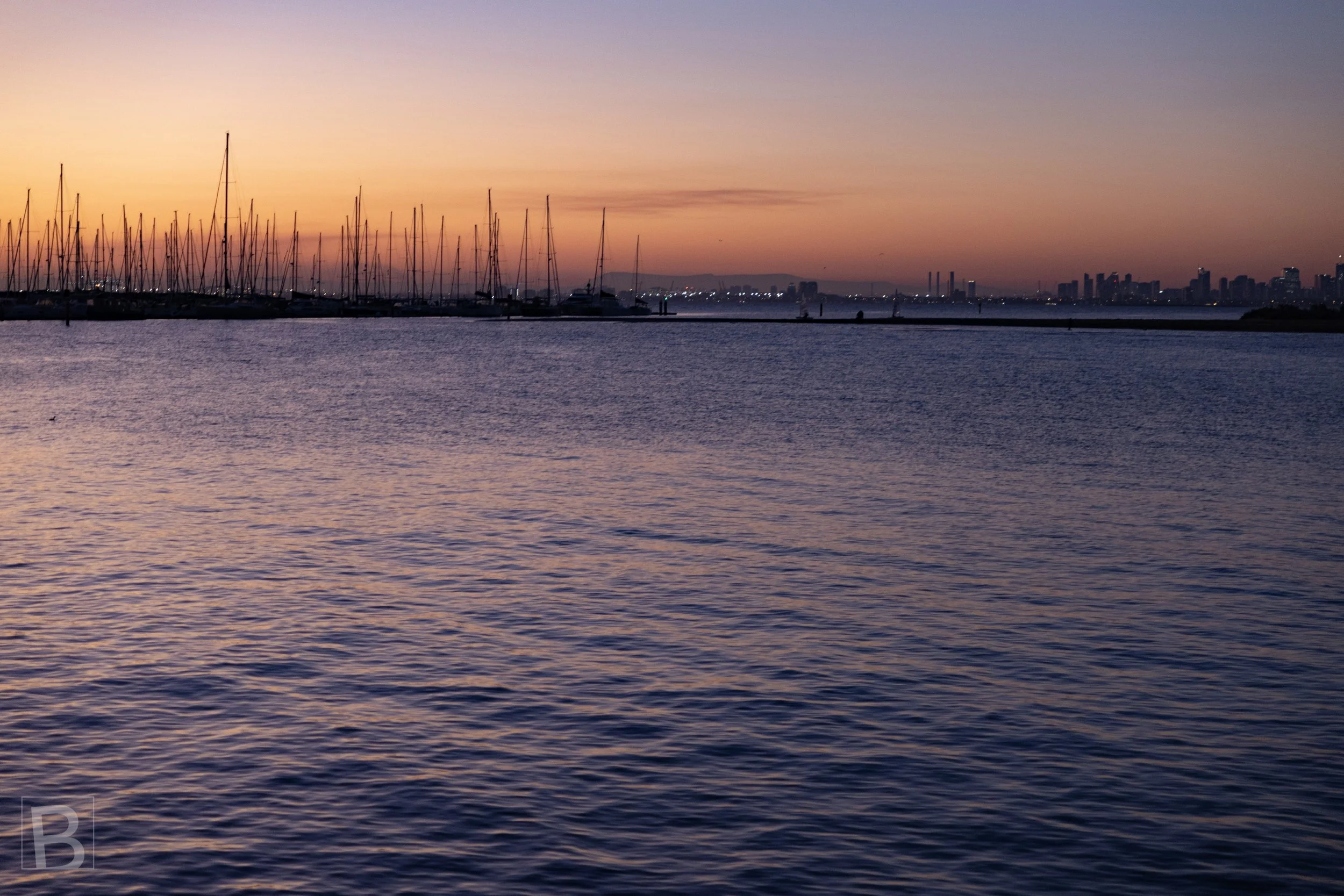 Fine masts gather along the horizon as sunset colour settles over a
quiet harbour and distant city skyline.